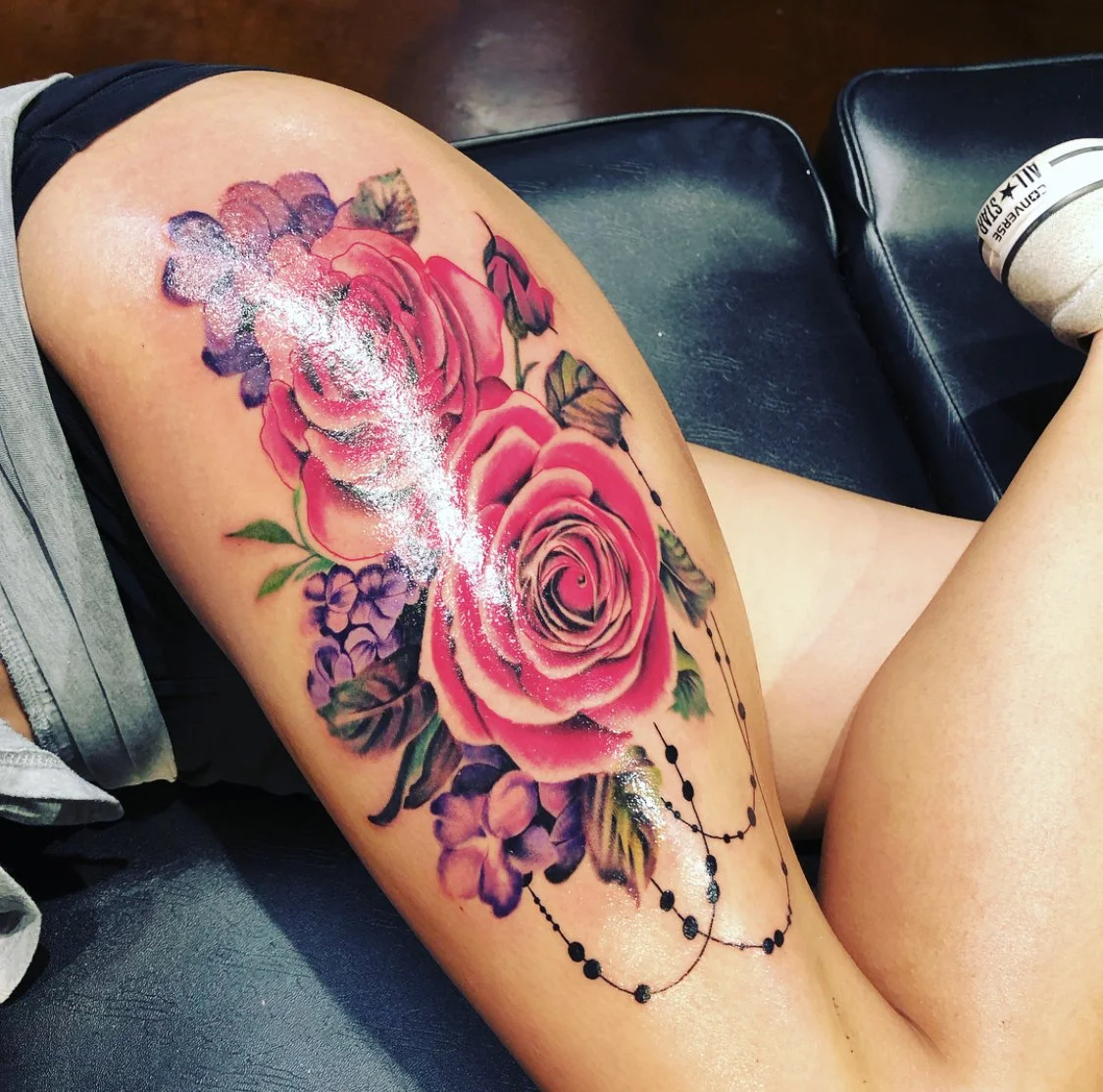 Colorful tattoo of pink roses and purple flowers with black beads on person’s thigh.