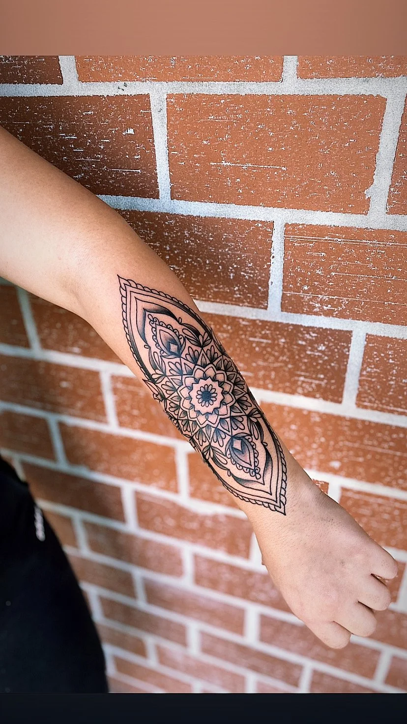 A person's forearm with a detailed black ink mandala tattoo, set against a brick wall background.