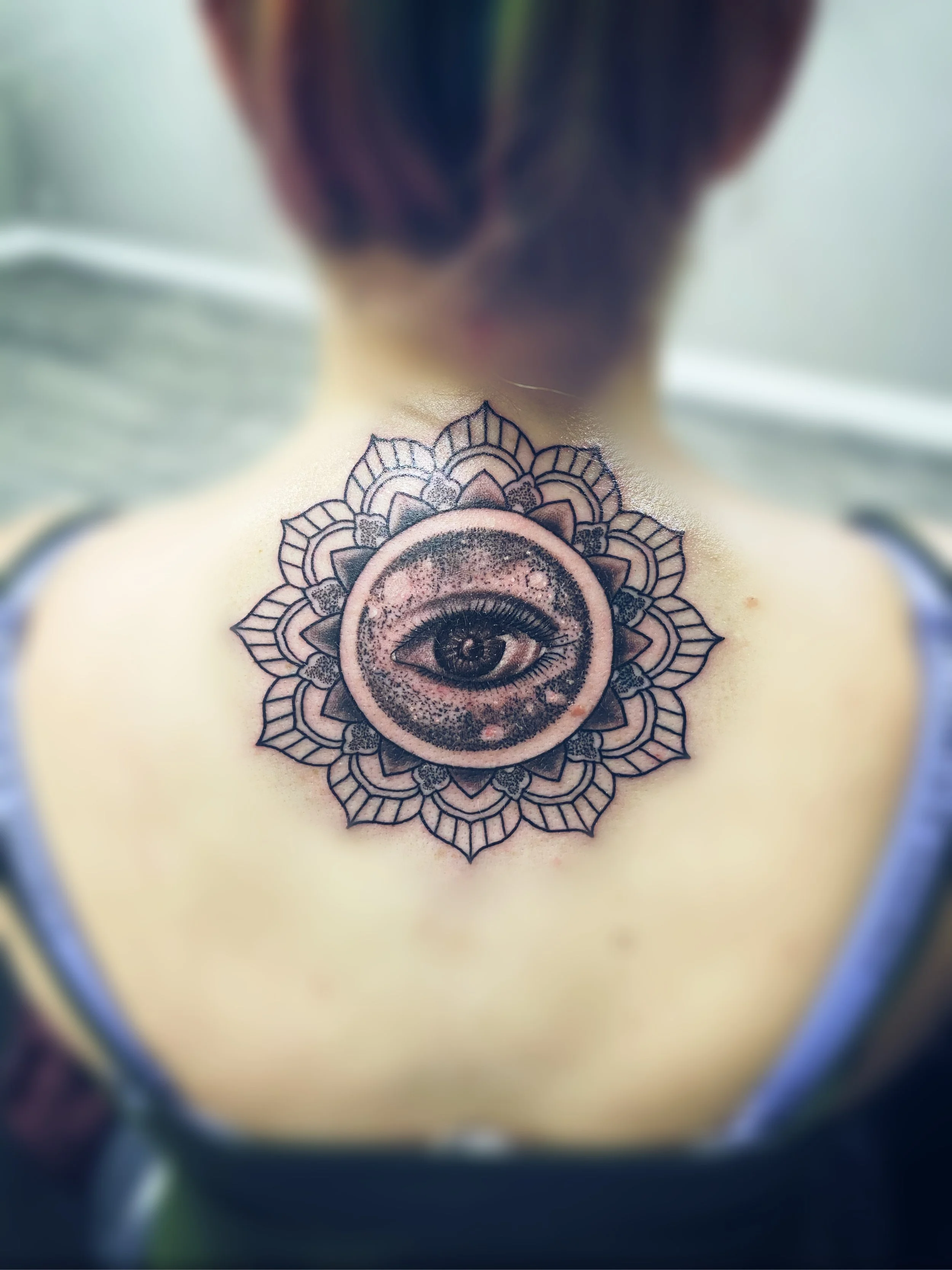 Tattoo of an eye within a mandala design on a person's chest, partially obscured by a blurry foreground.