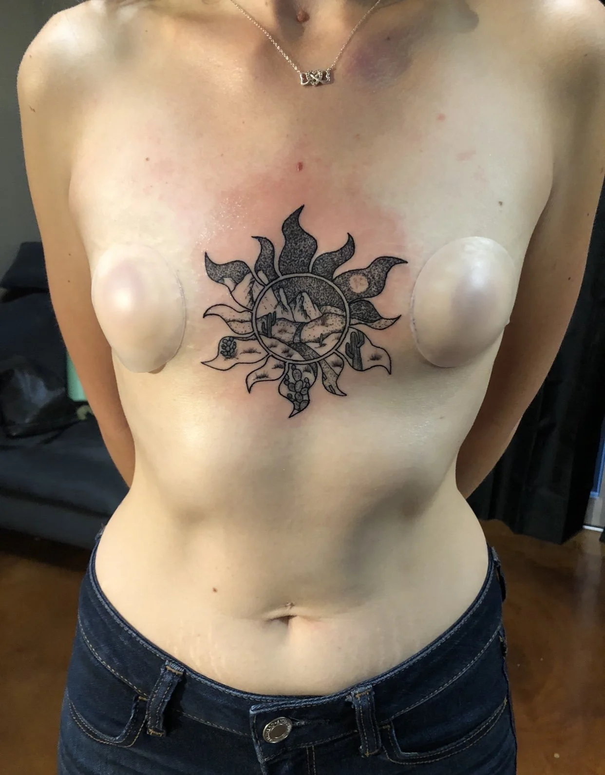 A woman with a tattoo of mountains, a lake, and a sun on her chest, and two prosthetic breasts.