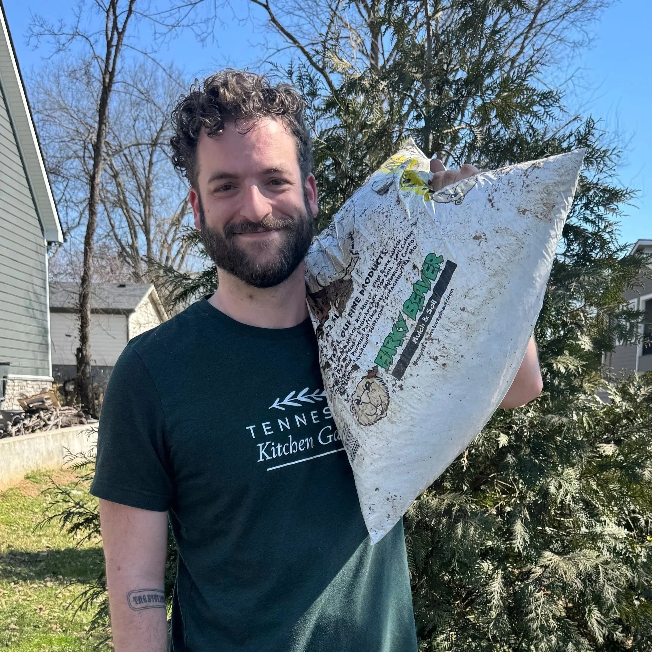 A smiling man with a beard and a beige cap outdoors near a house with blue siding and a tree.