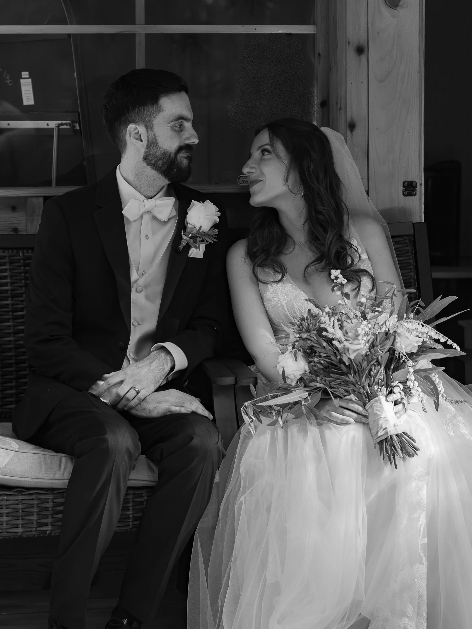 A black and white photo of a bride and groom sitting close together, gazing into each other's eyes. The groom is wearing a tuxedo with a bow tie and boutonniere, and the bride is in a wedding gown holding a bouquet of flowers. They are seated indoors