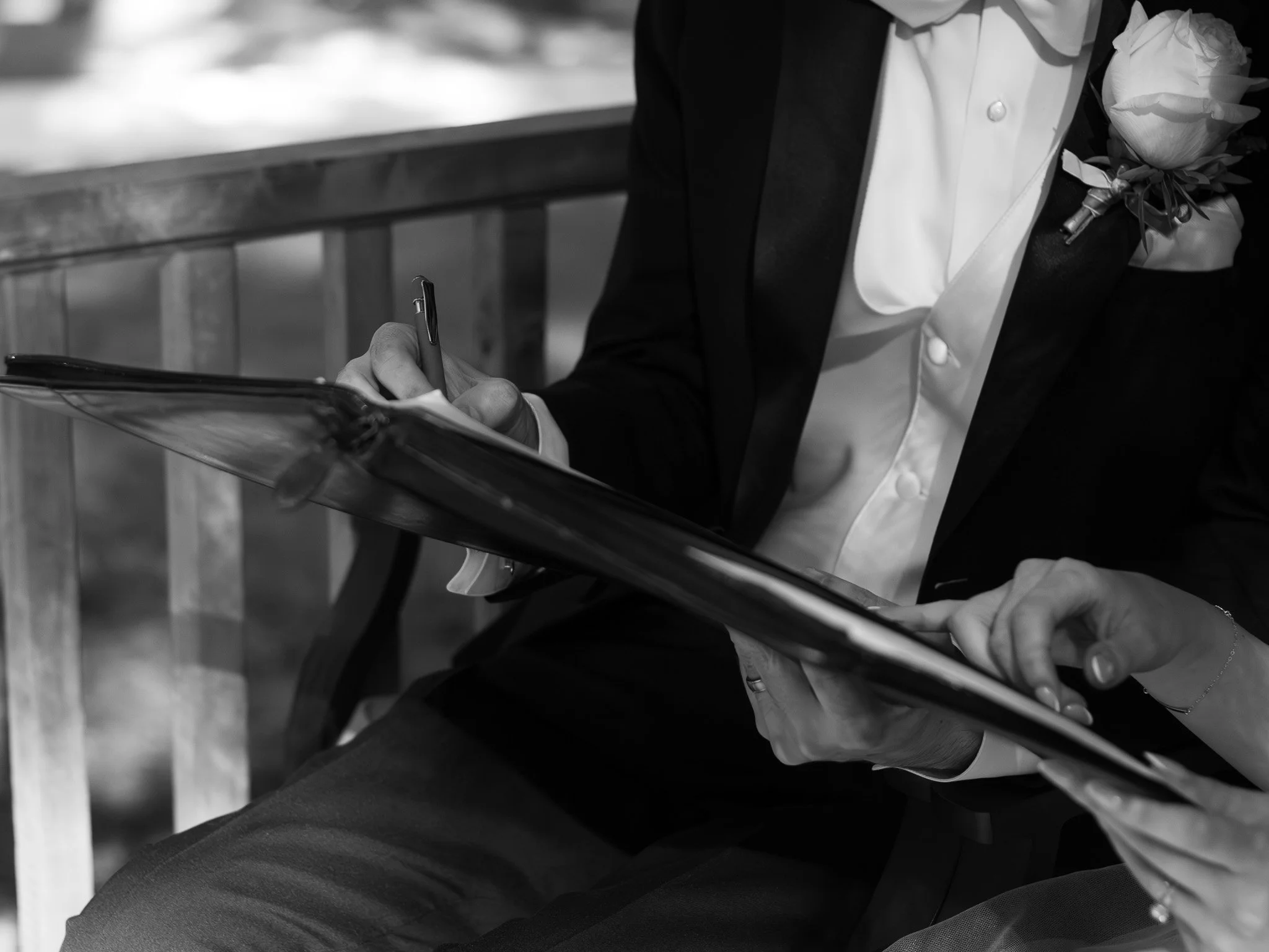 A person in formal attire sitting on a wooden bench, signing a document or guestbook with a pen, and wearing a boutonniere on their jacket.