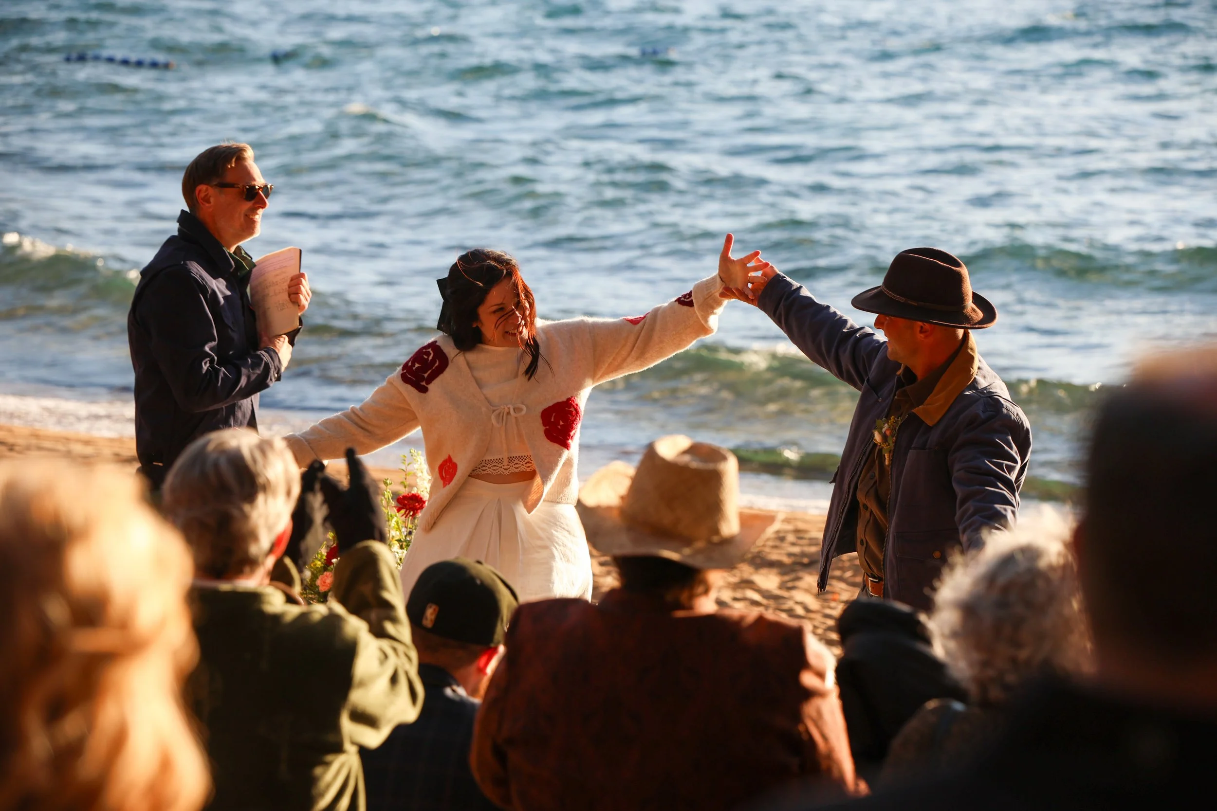 A beach wedding ceremony taking place at sunset, with a bride in a white outfit with red decorative patches, a groom in a dark jacket and hat, an officiant holding a book, and guests seated along the shoreline, ocean in the background.
