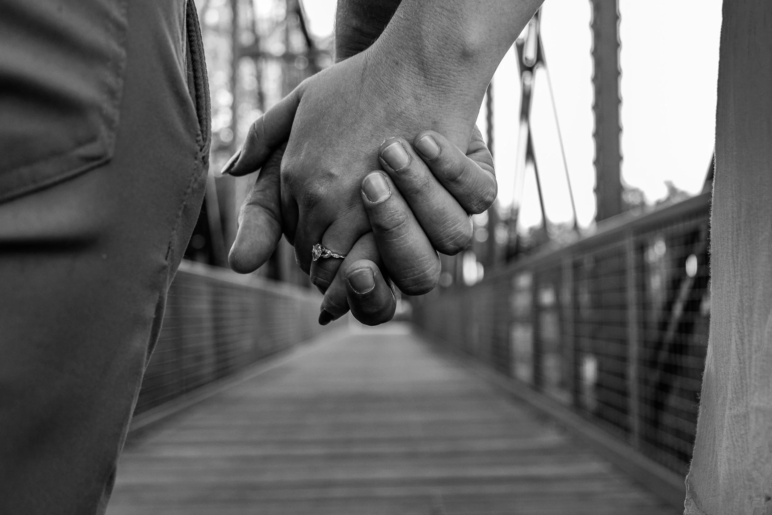 Close-up of a man and woman holding hands on a bridge, with the woman wearing a ring, black and white photo.