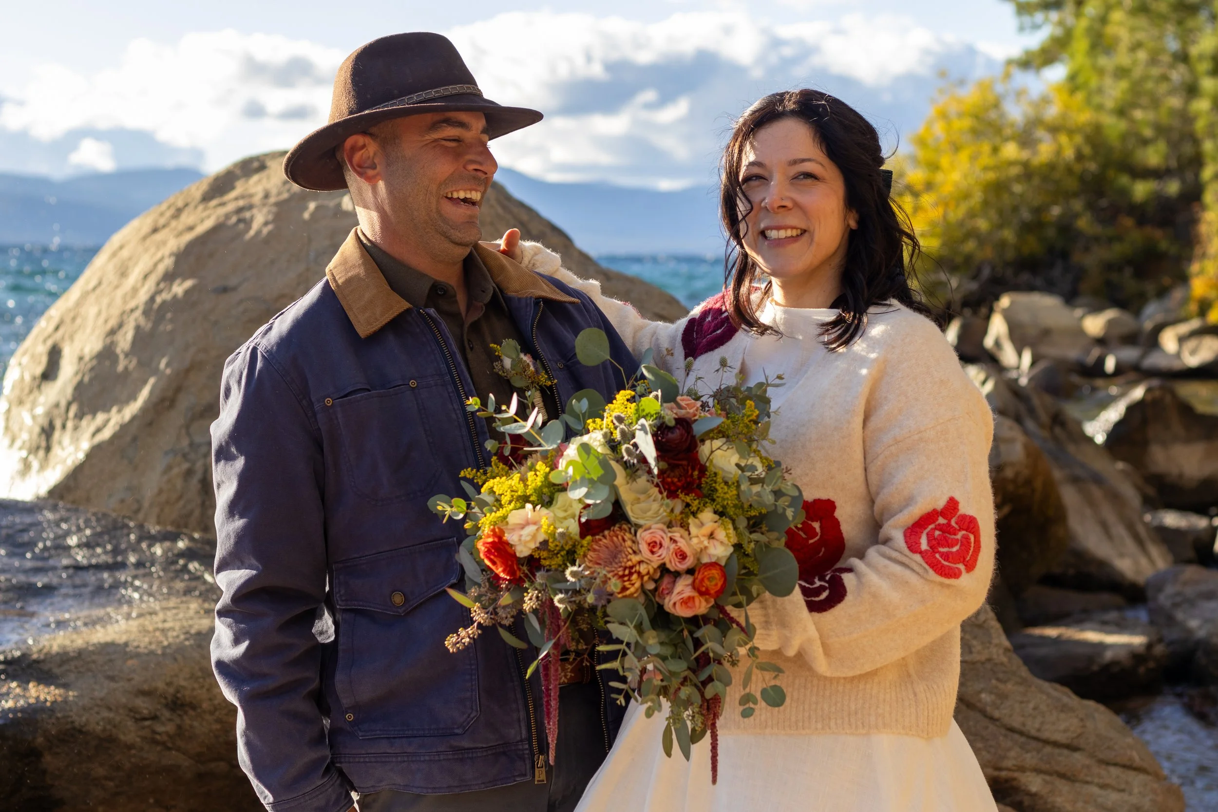 A couple stands outdoors near rocks and water, smiling and holding a bouquet of flowers. The man wears a dark hat and denim jacket, the woman wears a cream sweater with red floral embroidery. It appears to be during fall with trees showing autumn col
