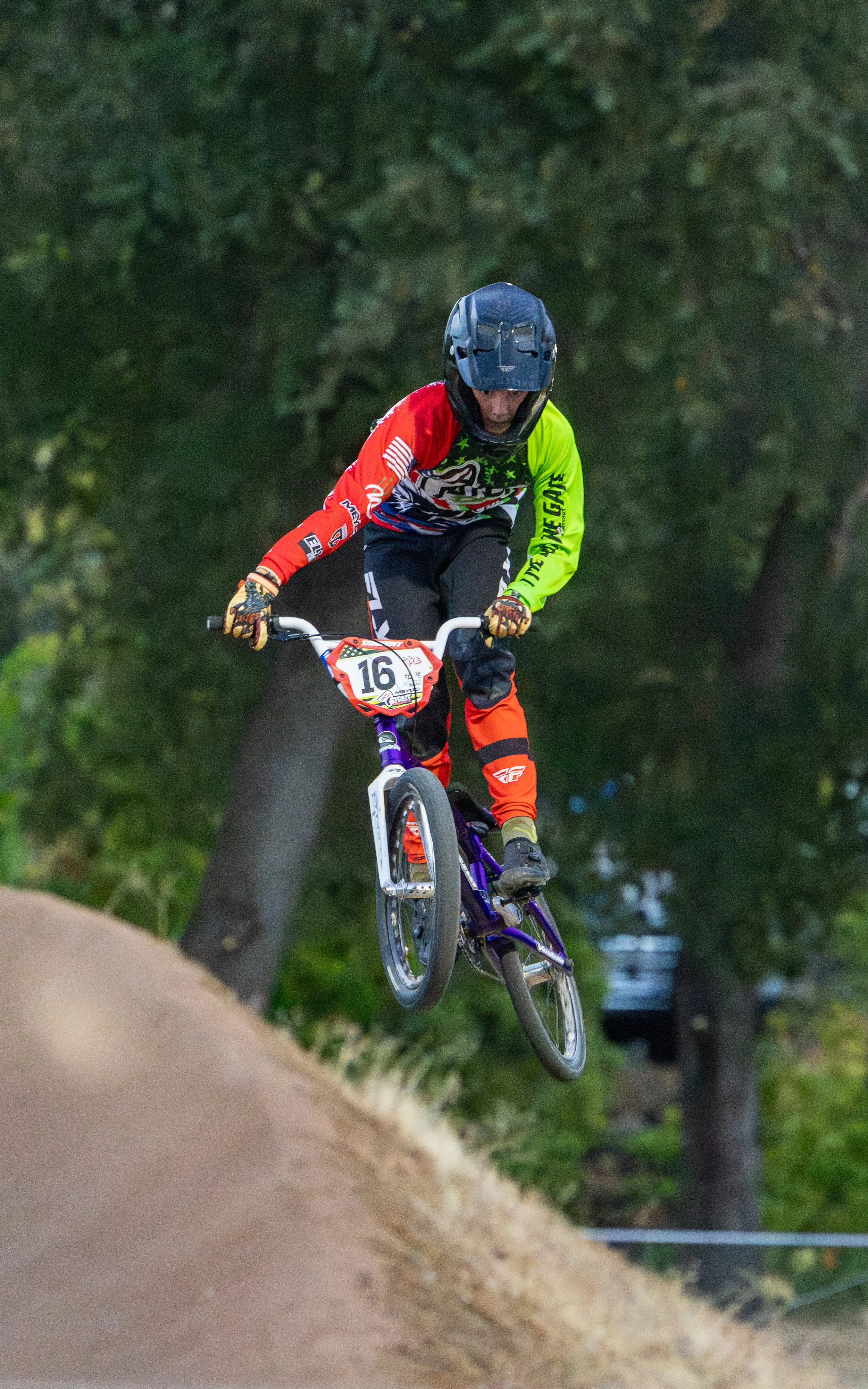 A young BMX rider in colorful gear performing a jump on a dirt track with trees in the background.