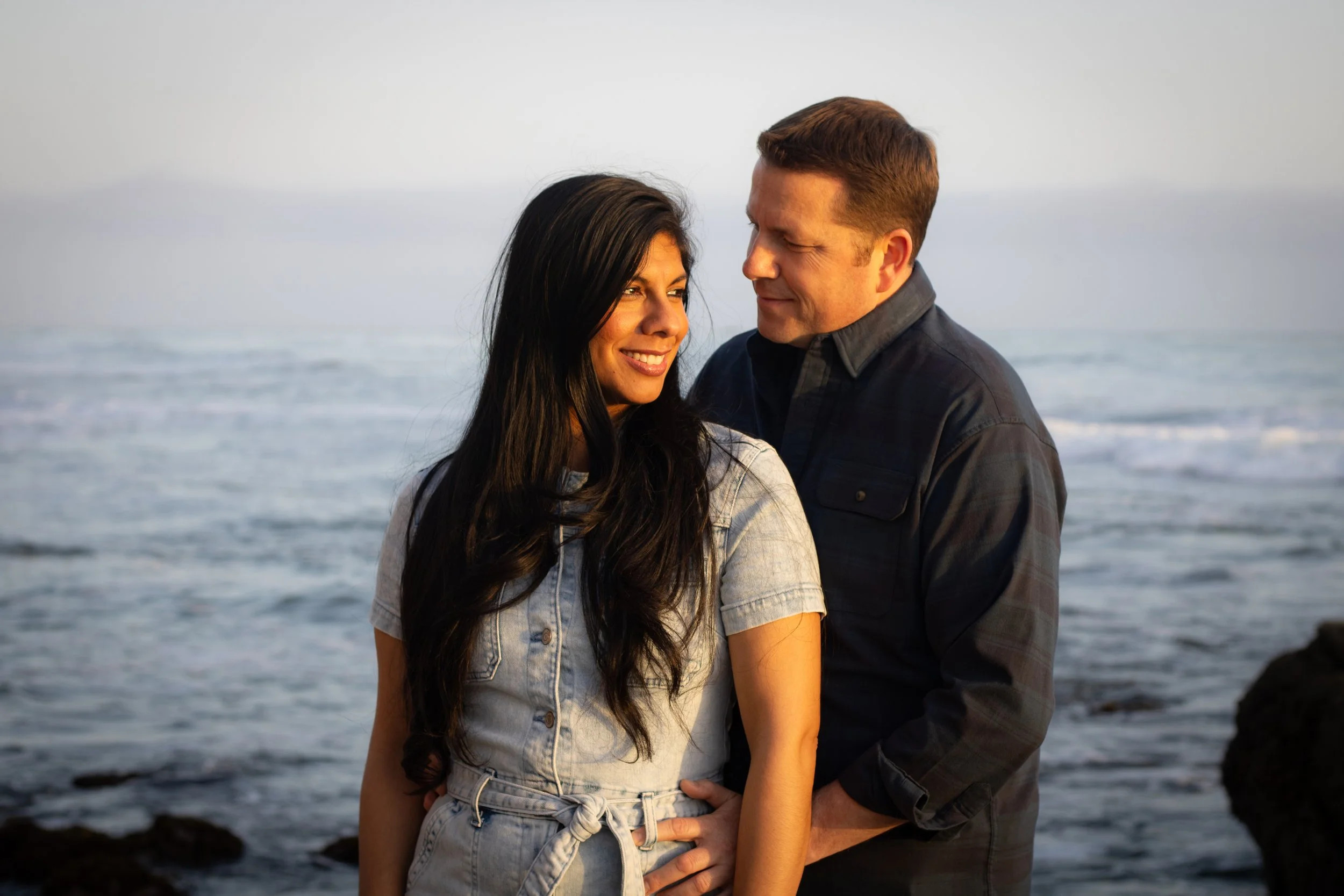 A couple on the beach during sunset, smiling and looking at each other, with the ocean in the background.