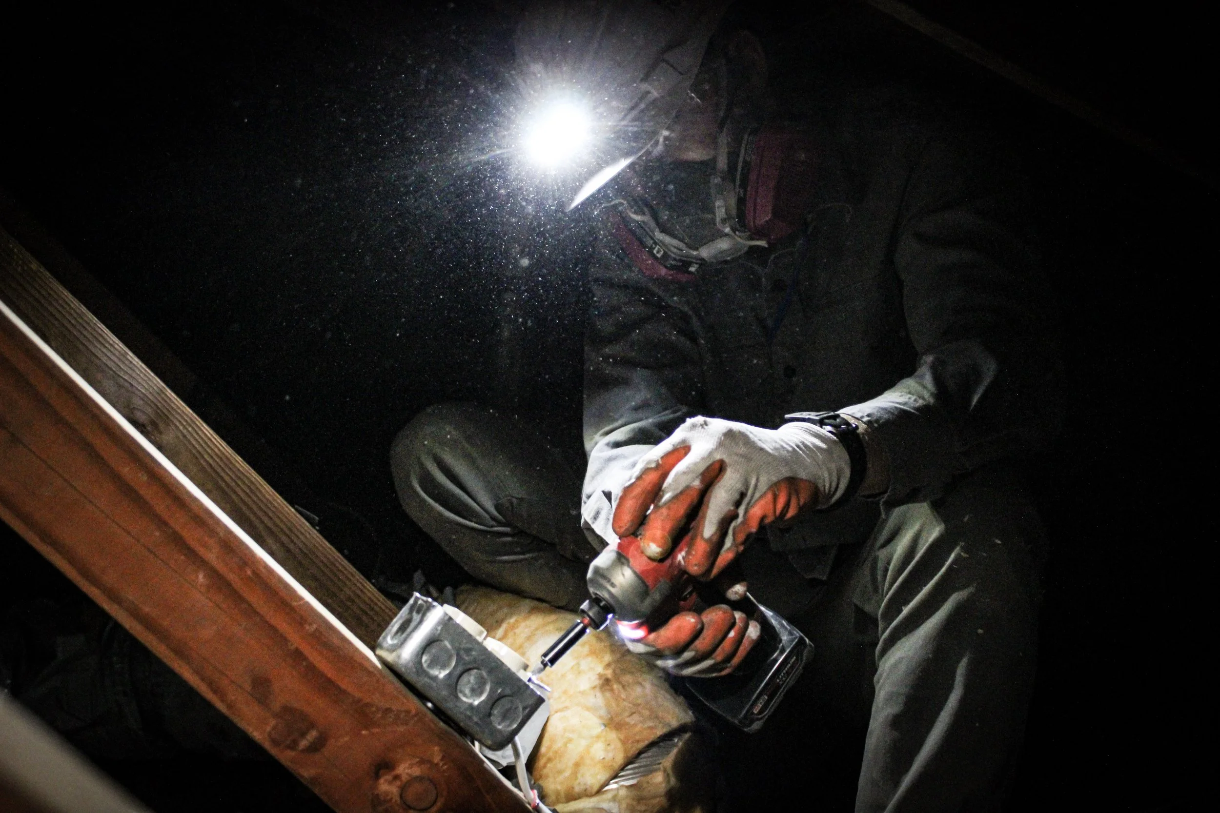 Person wearing gloves and safety gear working with a power tool in a dark space, illuminated by a bright light.