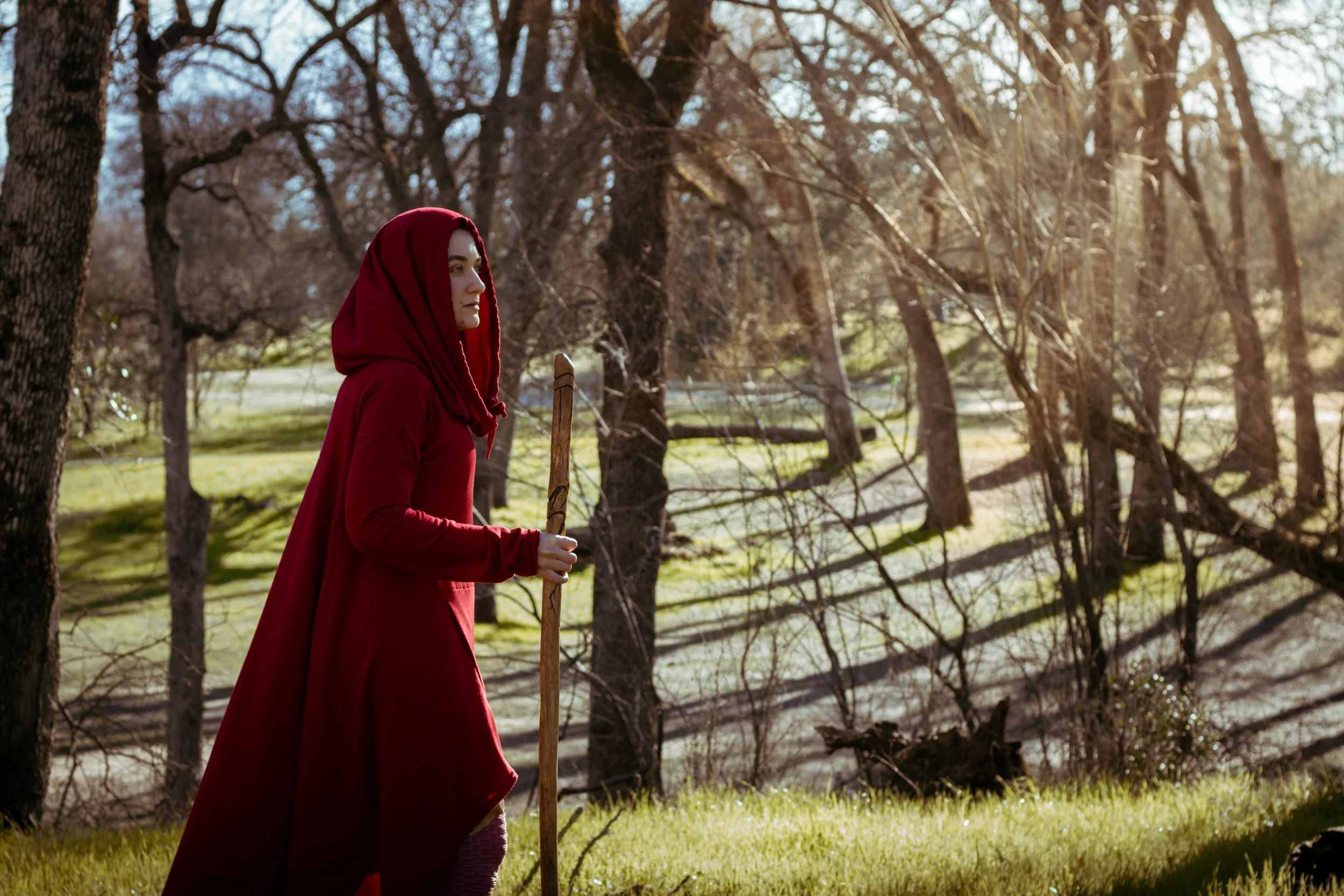A woman in a red hooded cloak walking through a sunlit wooded area with a wooden staff.