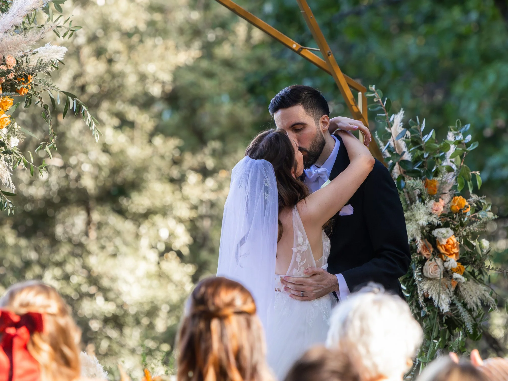 A bride and groom share a kiss during their outdoor wedding ceremony, surrounded by floral arrangements and greenery.