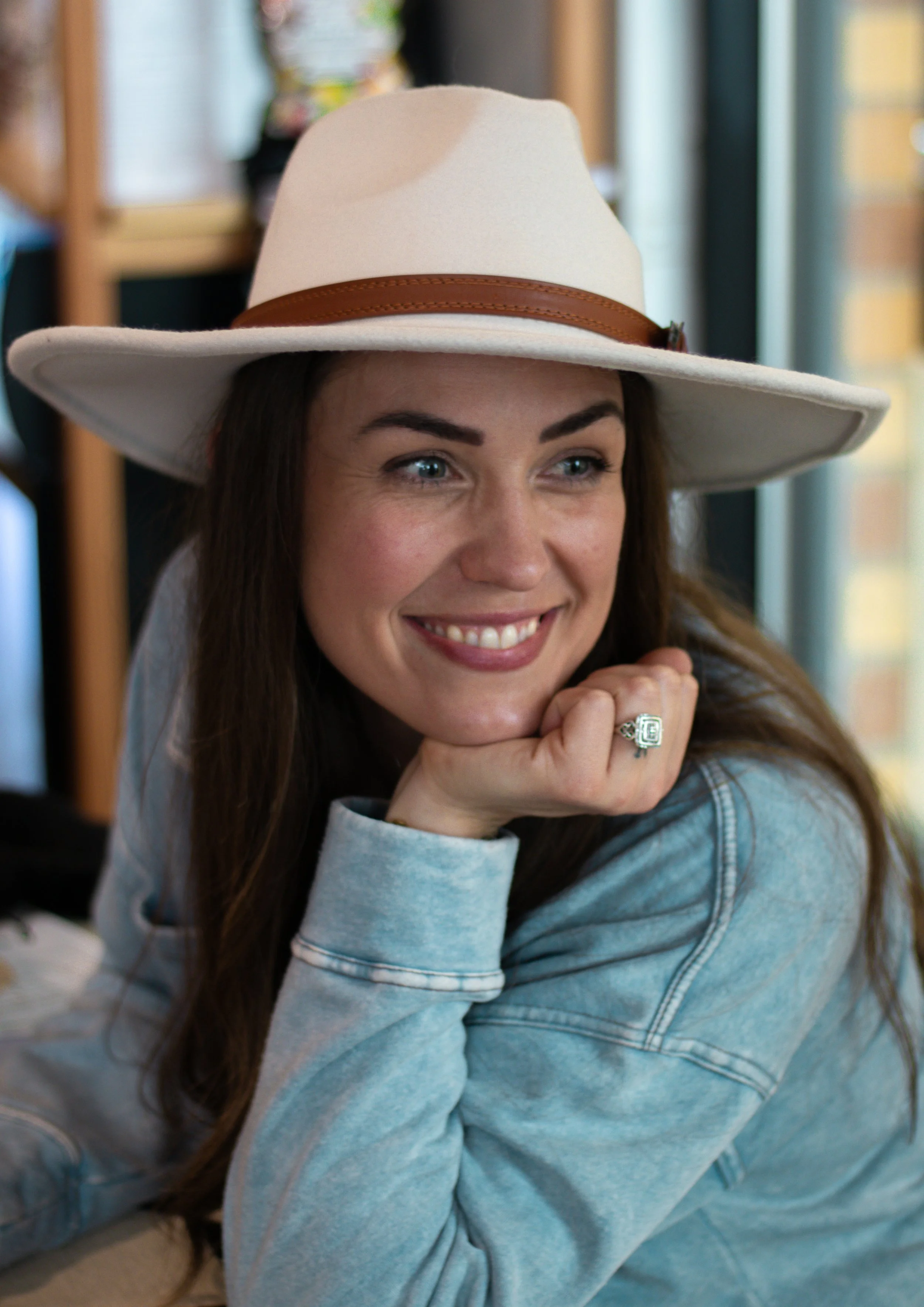 A woman with long brown hair wearing a large beige wide-brimmed hat with a brown band, smiling and resting her chin on her hand in a cozy indoor setting.