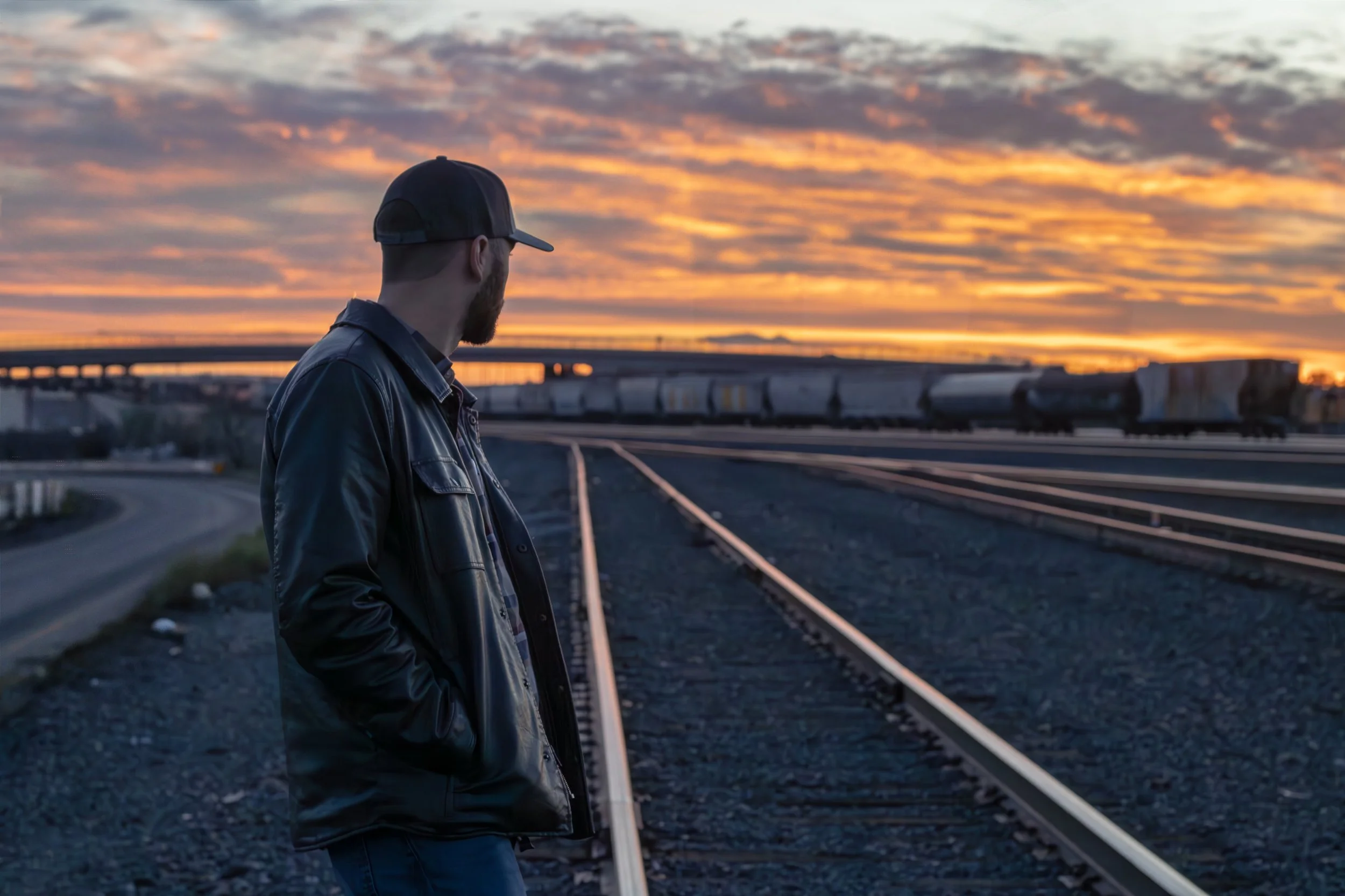 A man stands on railroad tracks at sunset, wearing a black leather jacket and baseball cap, looking towards the horizon with a train in the background.