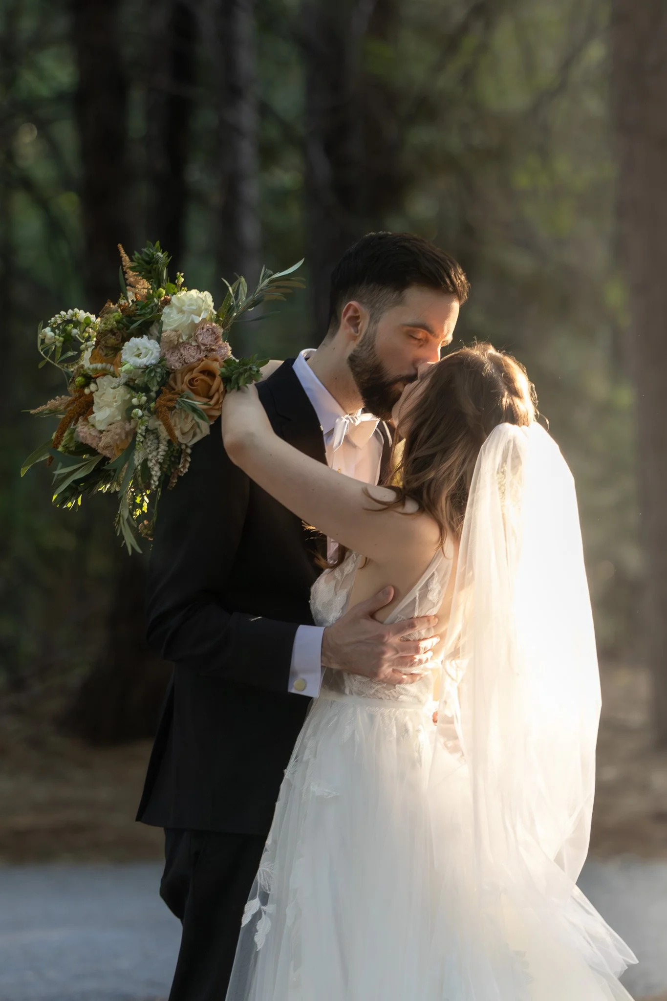 A newlywed couple sharing a kiss outdoors in a natural setting, with the bride holding a large bouquet of flowers and wearing a white wedding dress and veil.
