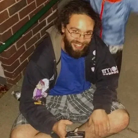 Man sitting on the ground outside next to a brick wall, wearing glasses, a hoodie, blue shirt, plaid shorts, and sneakers.