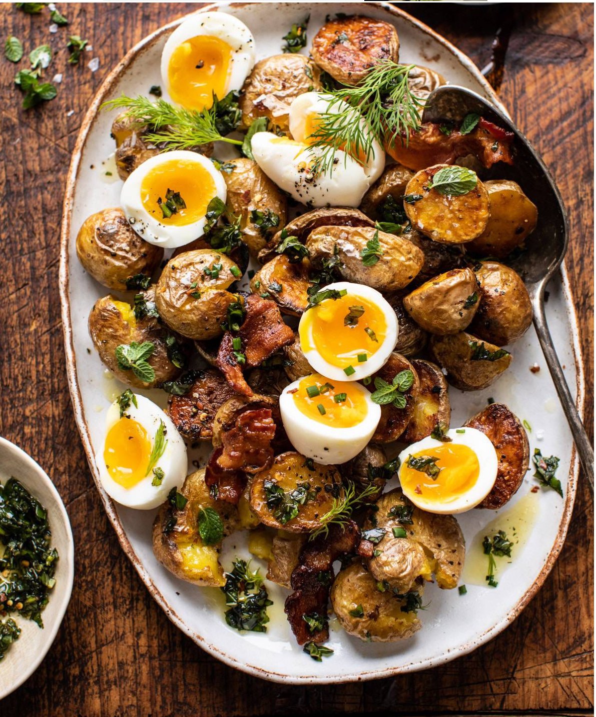 Roasted potatoes and soft-boiled eggs garnished with herbs and bacon on a white oval plate, wooden table.