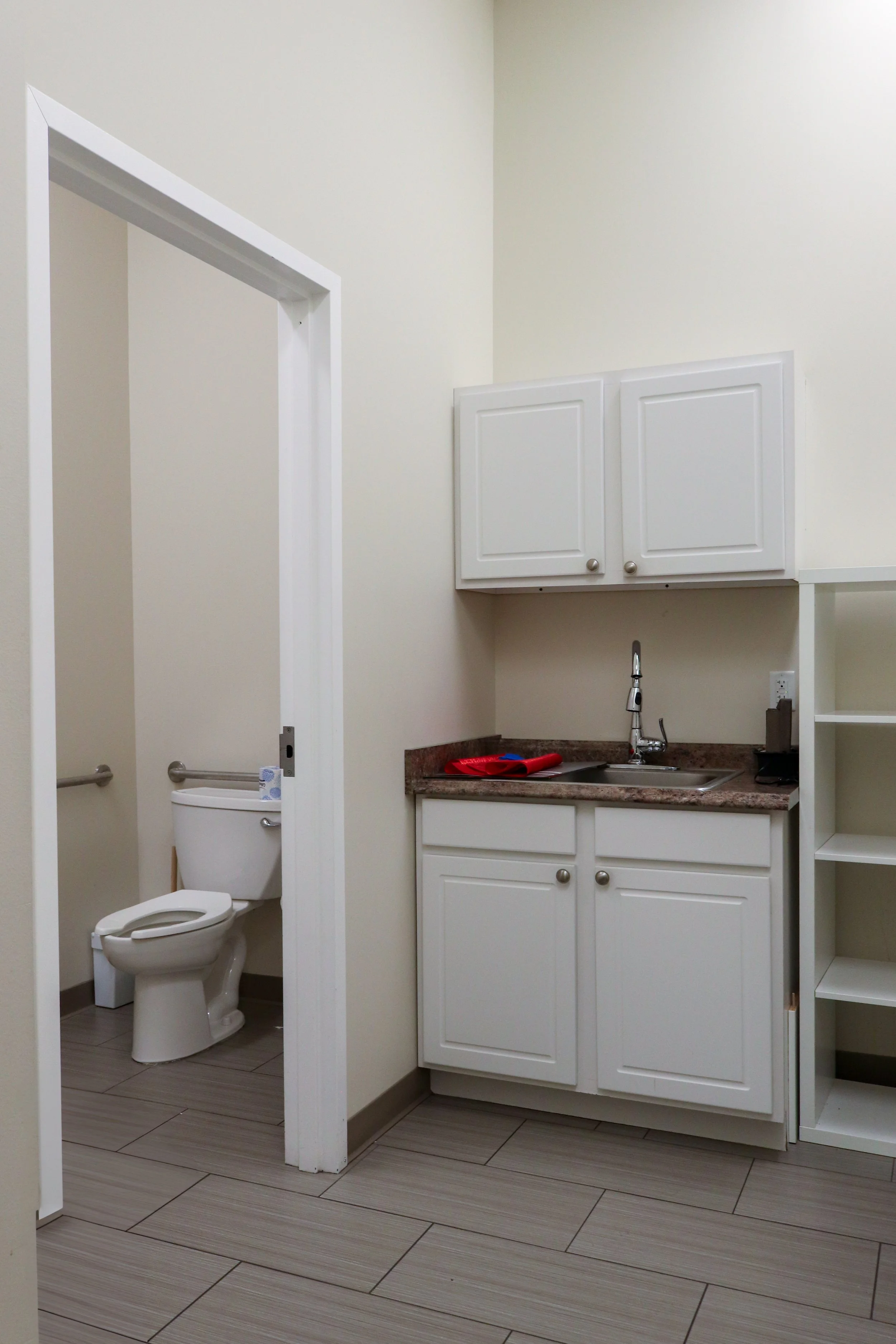 Bathroom interior with open door showing toilet, sink with faucet, white cabinets, and tiled floor.