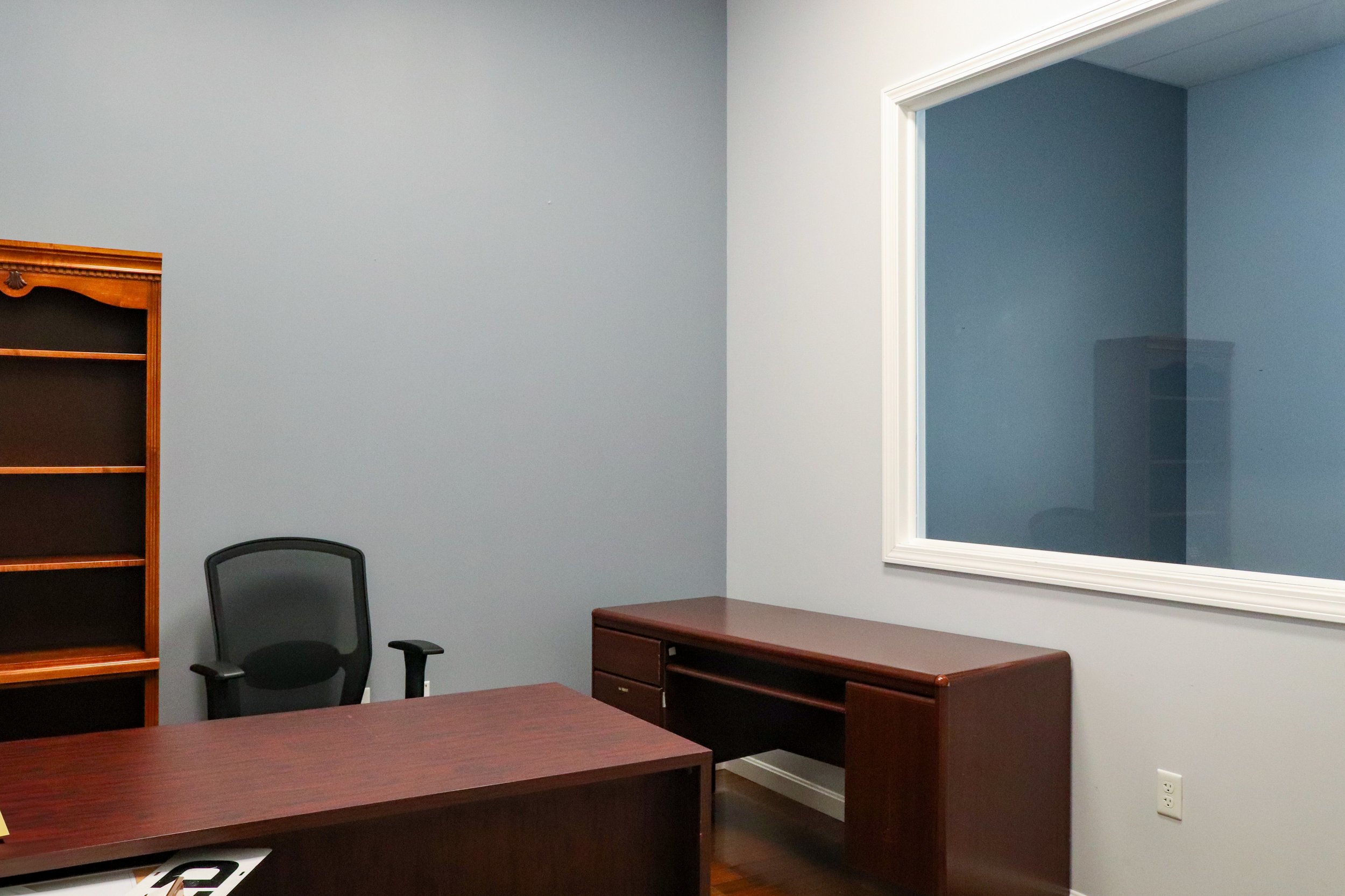 Empty office with wooden desk and chair, wooden bookcase, and a large window in a blue-walled room.