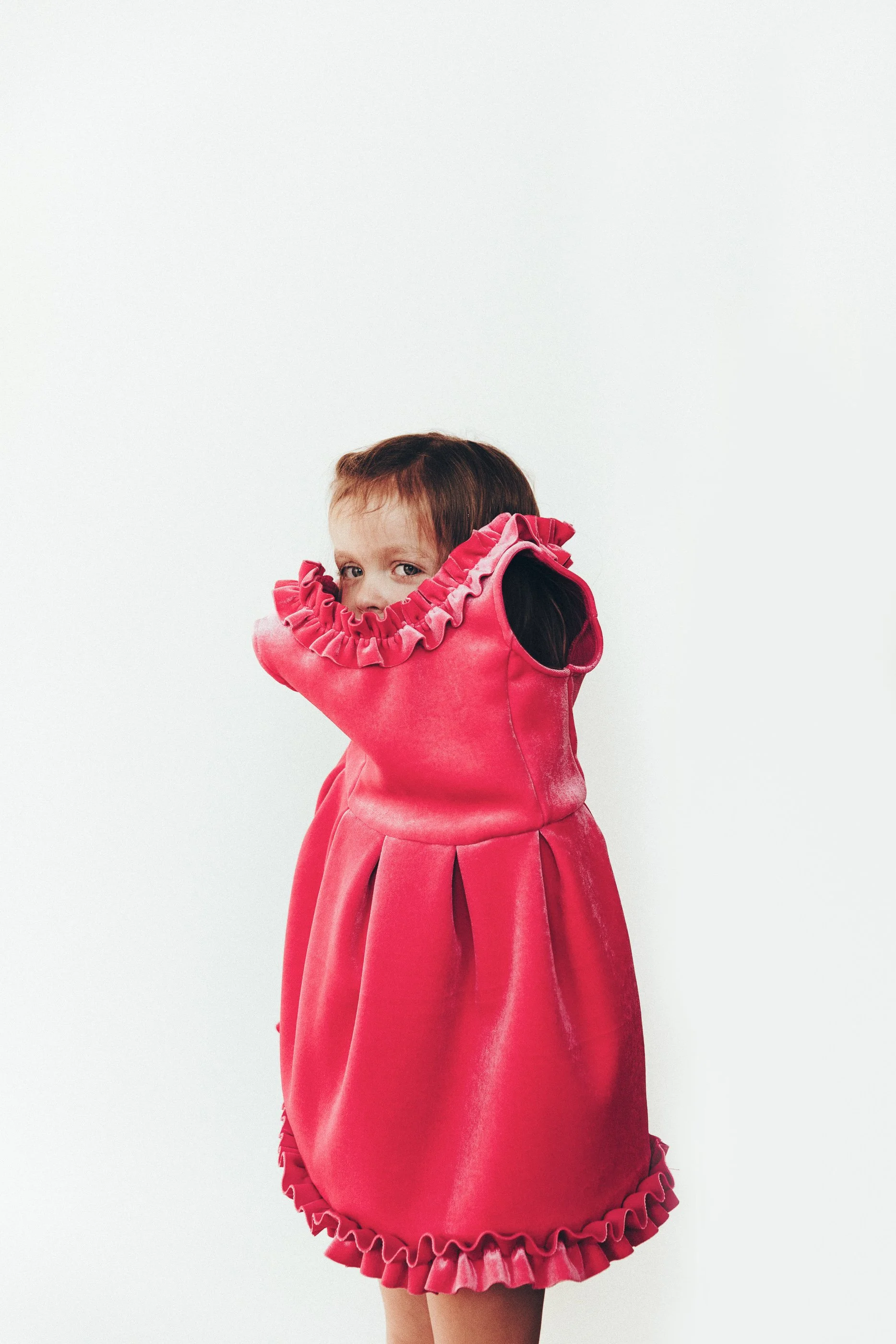 A young girl with brown hair wearing a pink dress with ruffled edges around the collar and hem, covering her face with her arm and looking over her shoulder.