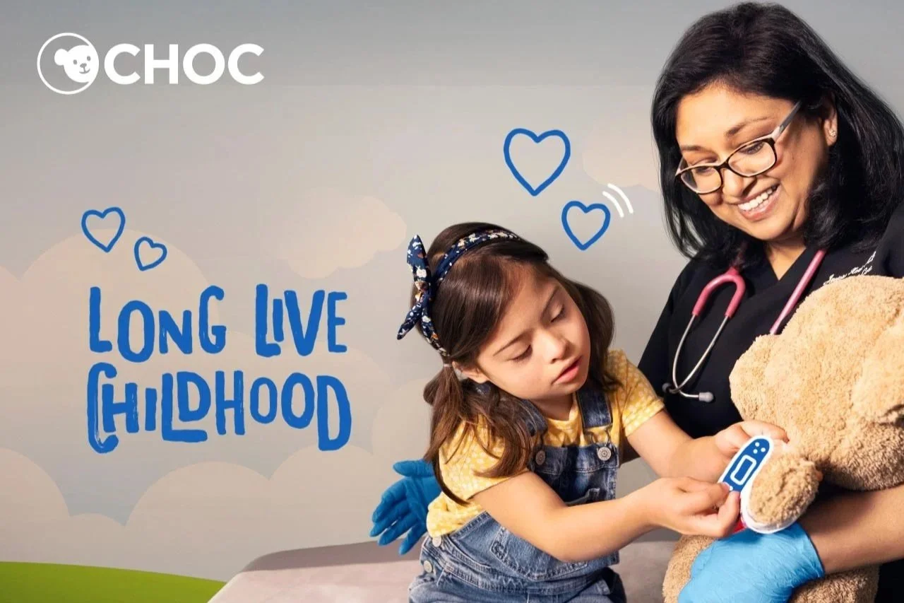 A young girl with a headband holding a thermometer to a teddy bear's paw while a smiling woman, possibly a nurse or doctor, helps her. The background has clouds, hearts, and the text 'Long Live Childhood' with a logo in the top left corner.