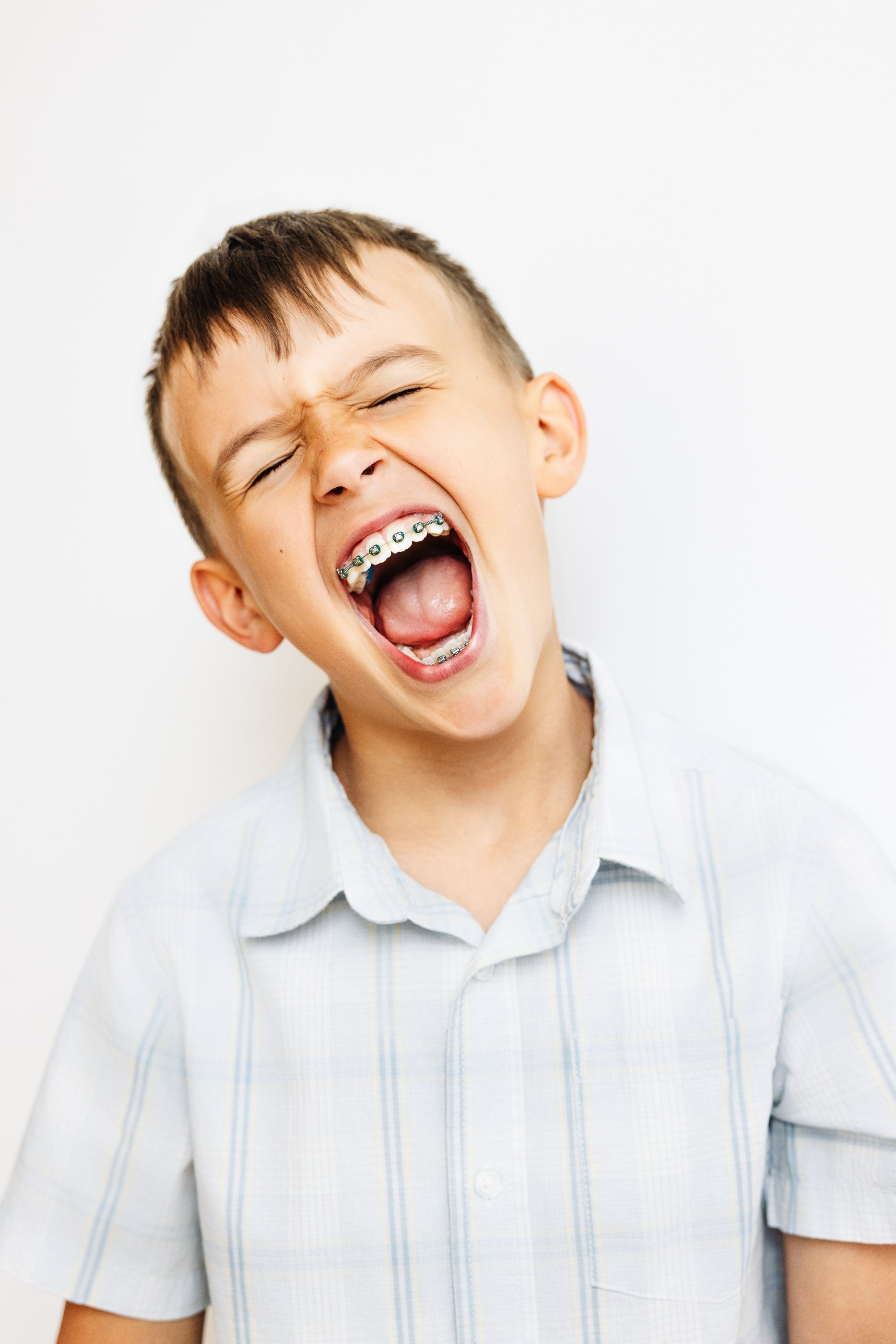 A young boy with orthodontic braces on his teeth, closing his eyes and smiling widely against a plain white background.