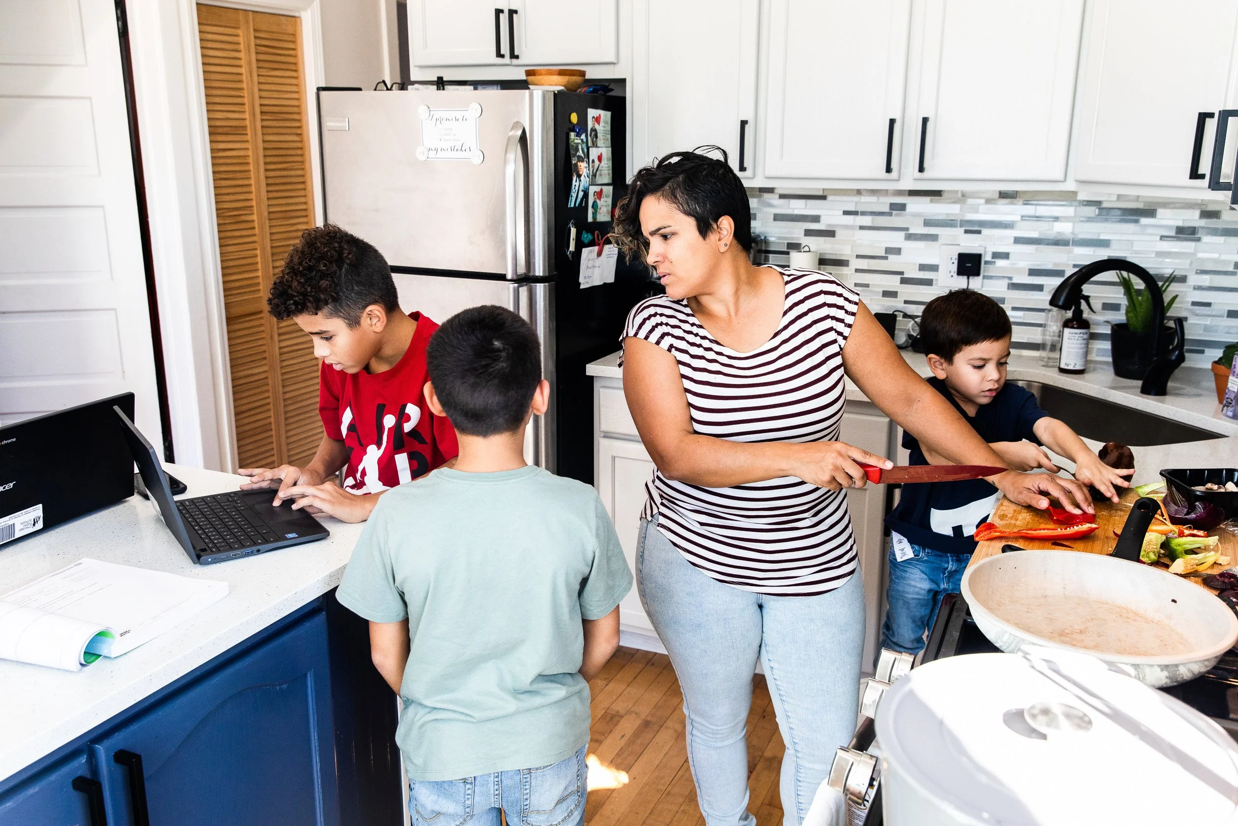 A woman chopping vegetables in a kitchen with three boys nearby, one using a laptop and another looking at a tablet.