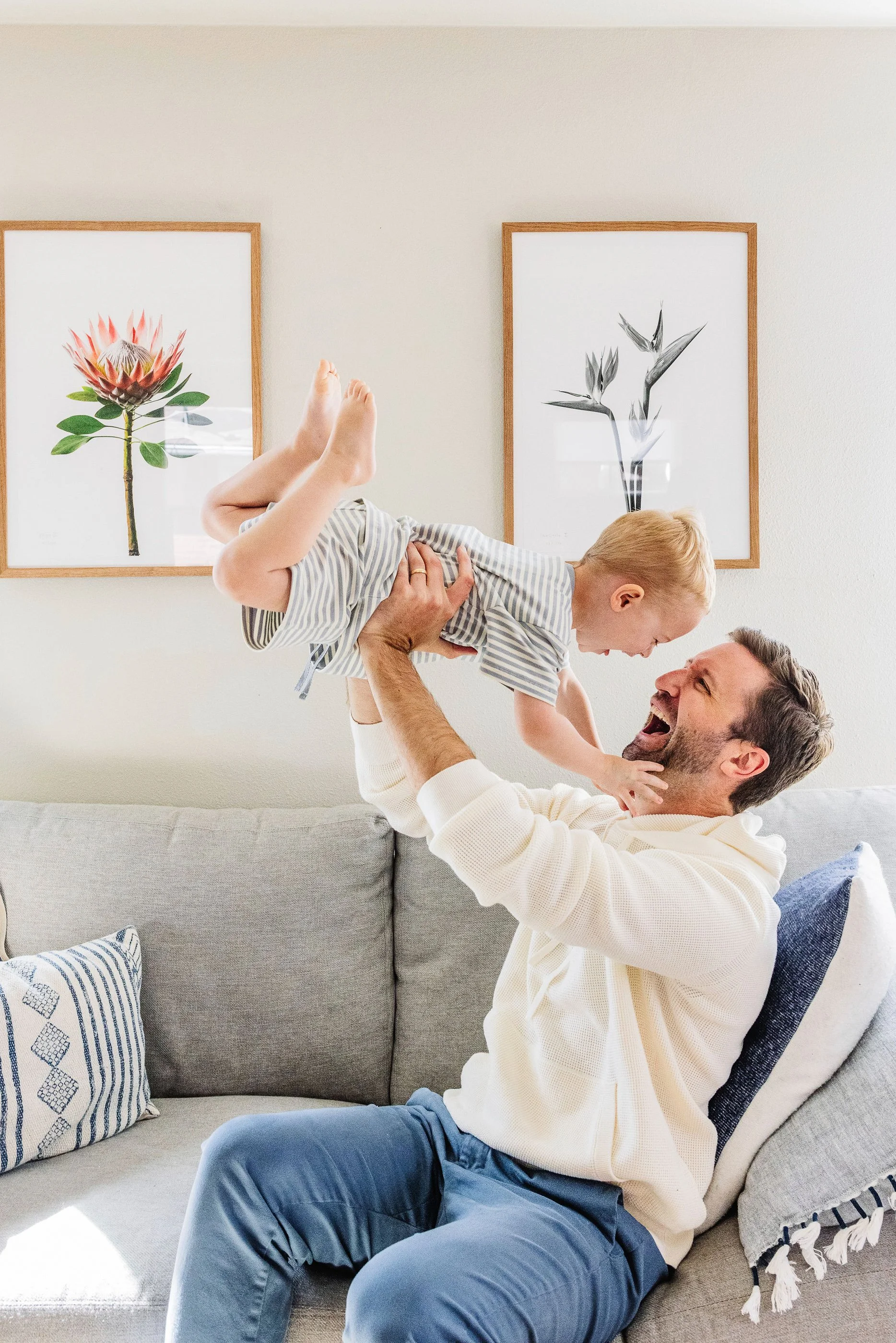 A man sitting on a beige couch lifting a young child with blonde hair and striped shirt over his head. The man has a beard and is wearing a cream-colored sweater, smiling as he interacts with the child. Two framed flower art prints hang on the white 