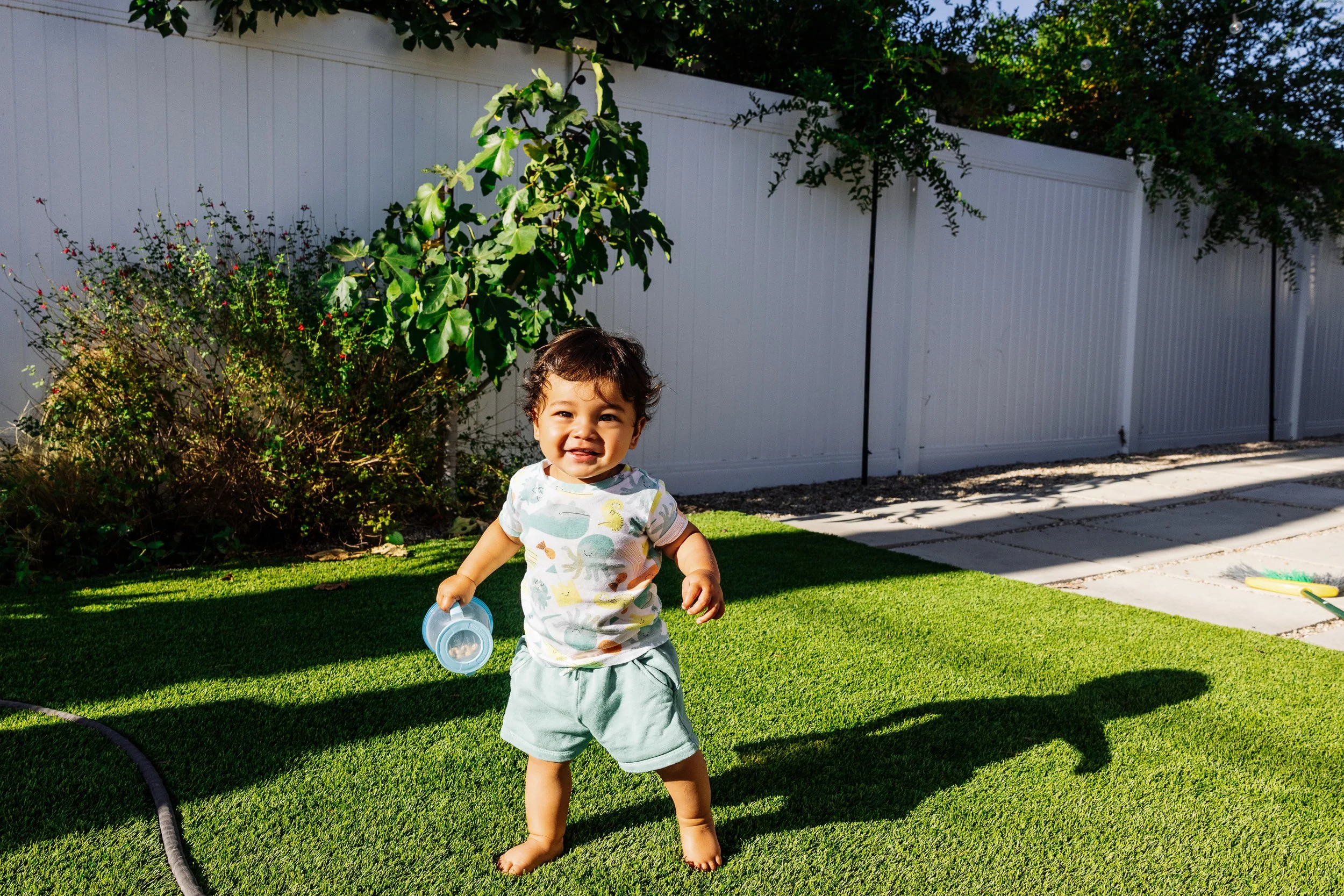 A young child with curly hair smiling while walking barefoot on grass, holding a blue container, outdoors in a backyard with a white fence and green trees nearby.