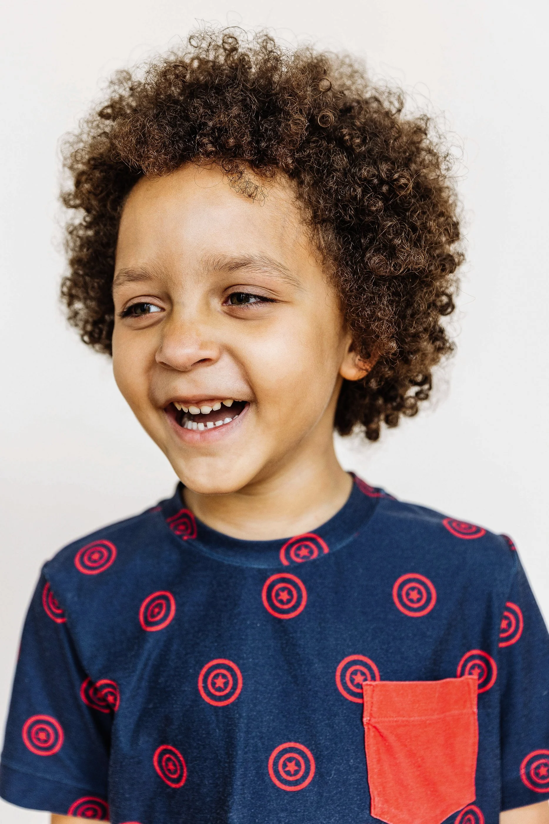 A young boy with curly brown hair smiling and looking to the side, wearing a navy blue shirt with red target patterns and a red pocket