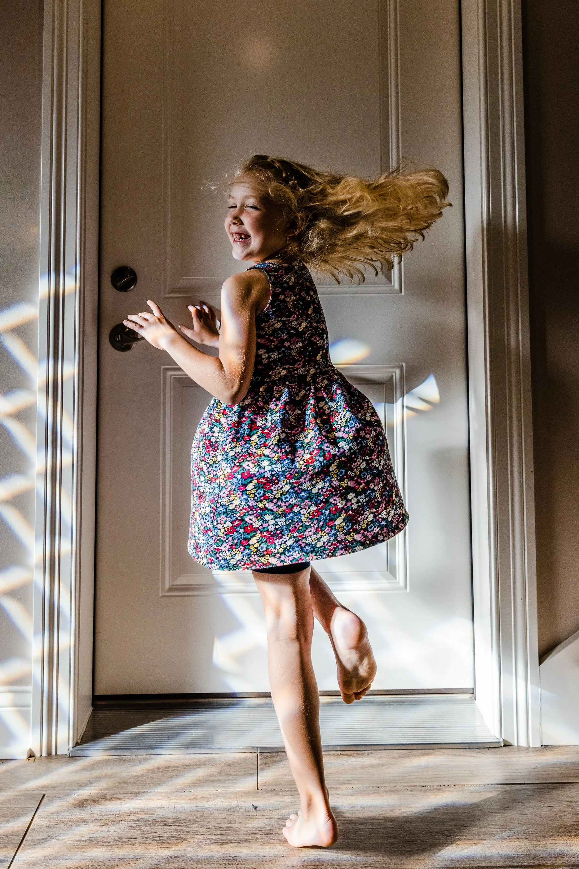 A young girl with long, flowing hair is spinning by a door, smiling and kicking one leg up in the air. She is wearing a sleeveless, floral-patterned dress and is barefoot. Sunlight creates rainbow light patterns on the floor and door.