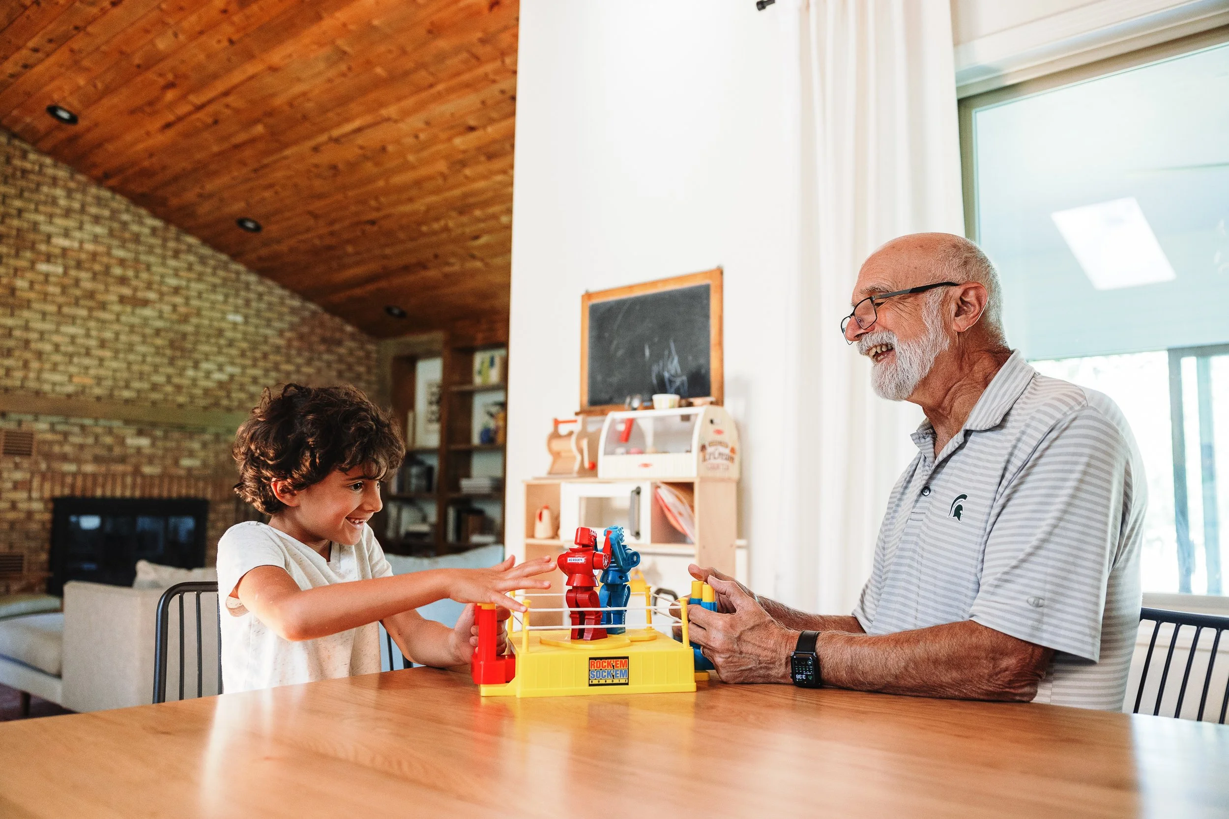 An elderly man with glasses and a white beard playing a board game with a young girl with curly dark hair at a wooden table in a bright, cozy room with brick and wood walls.