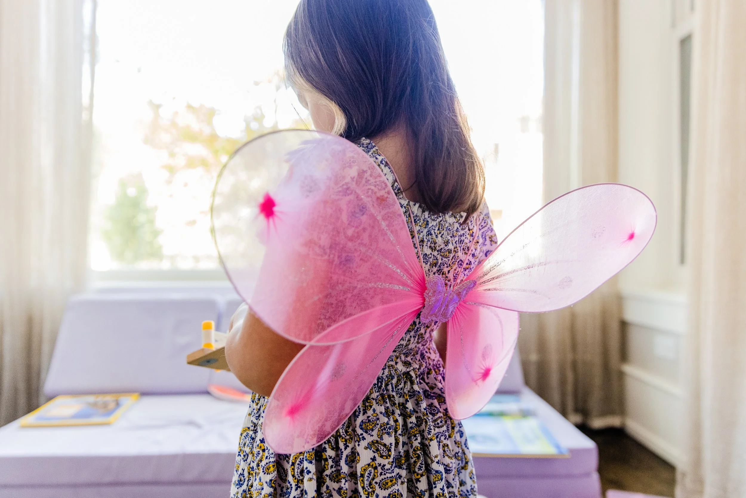 Child with pink fairy wings wearing a patterned dress, standing indoors near a table with books and papers, back facing camera.