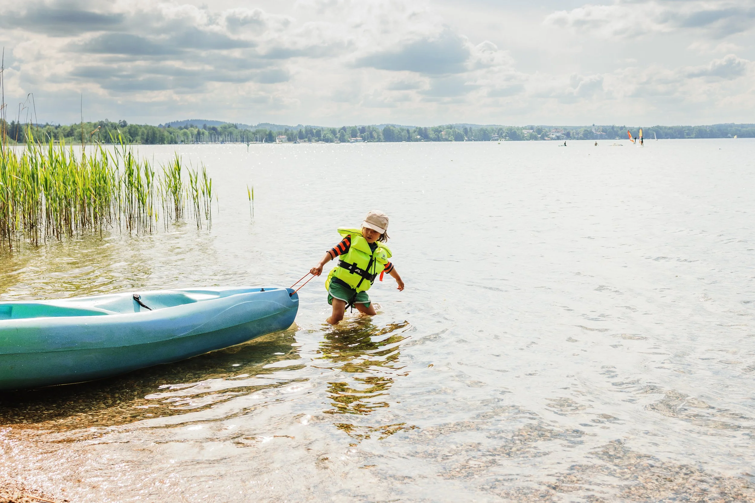 Child in a life jacket pulling a kayak out of the water at the lake shore on a cloudy day.