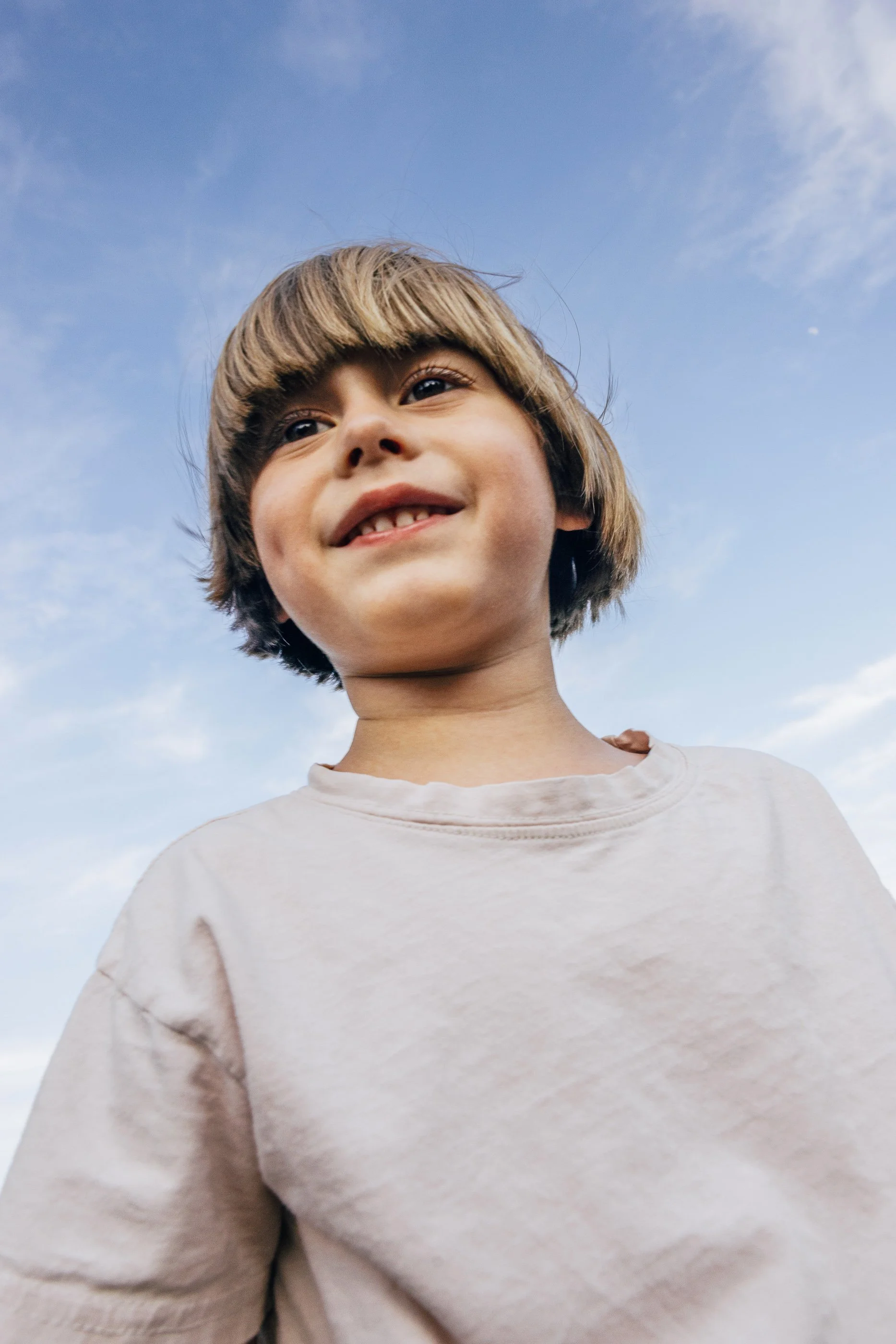 Close-up of a young boy with light brown hair smiling outdoors against a blue sky with wispy clouds.