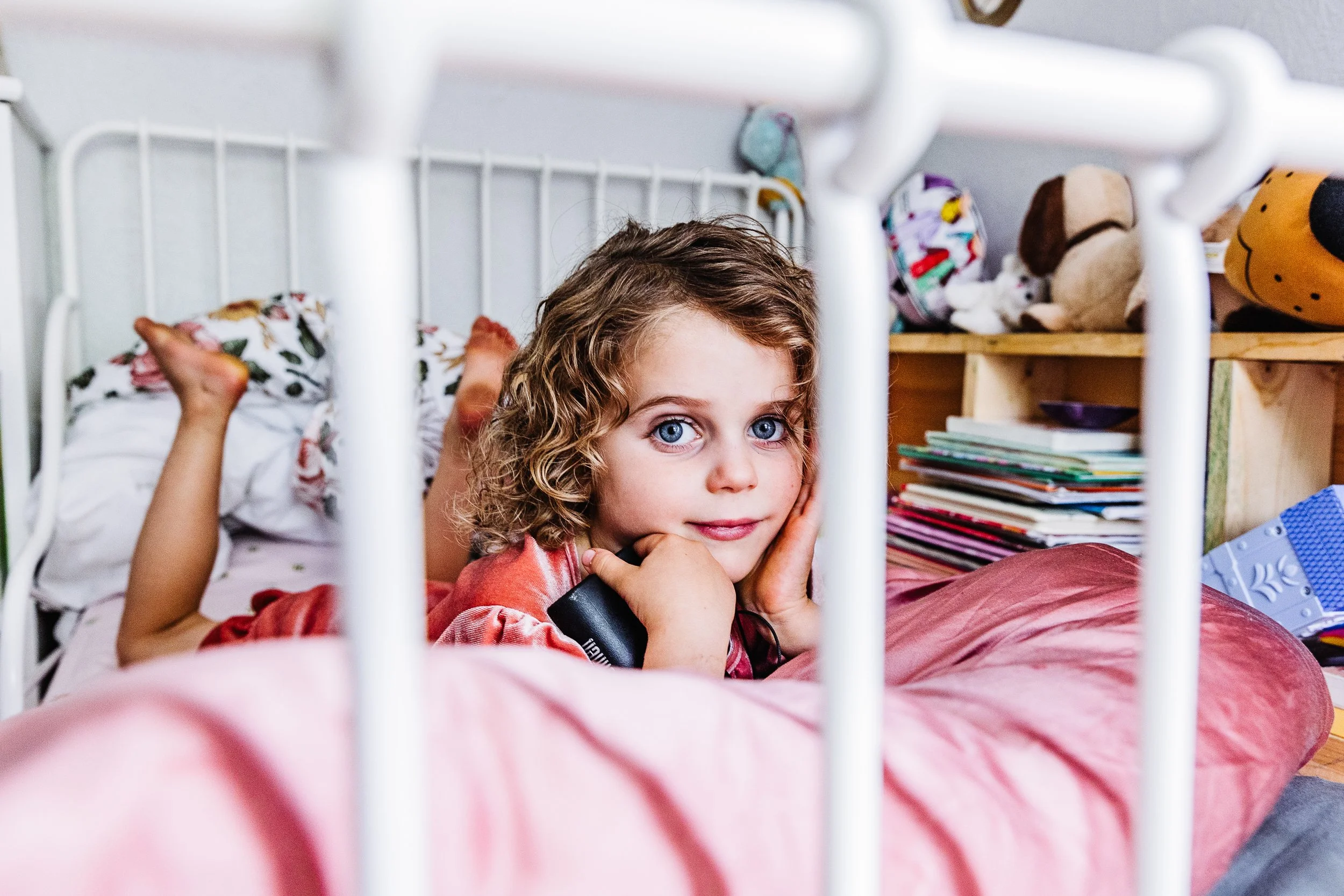 A young girl with curly blonde hair and blue eyes lying in a bed, resting her head on her hand, with a thoughtful expression, surrounded by toys and books in the background.