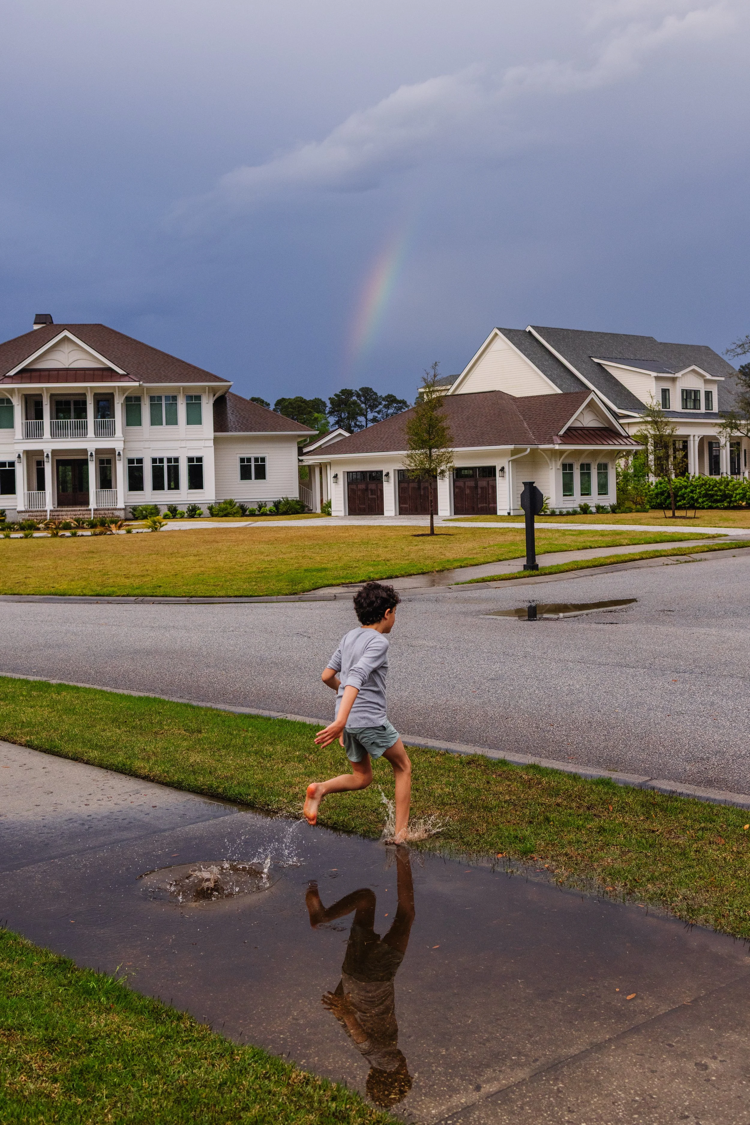 A young boy runs through a puddle on a suburban street with houses and trees, under a cloudy sky with a rainbow visible.