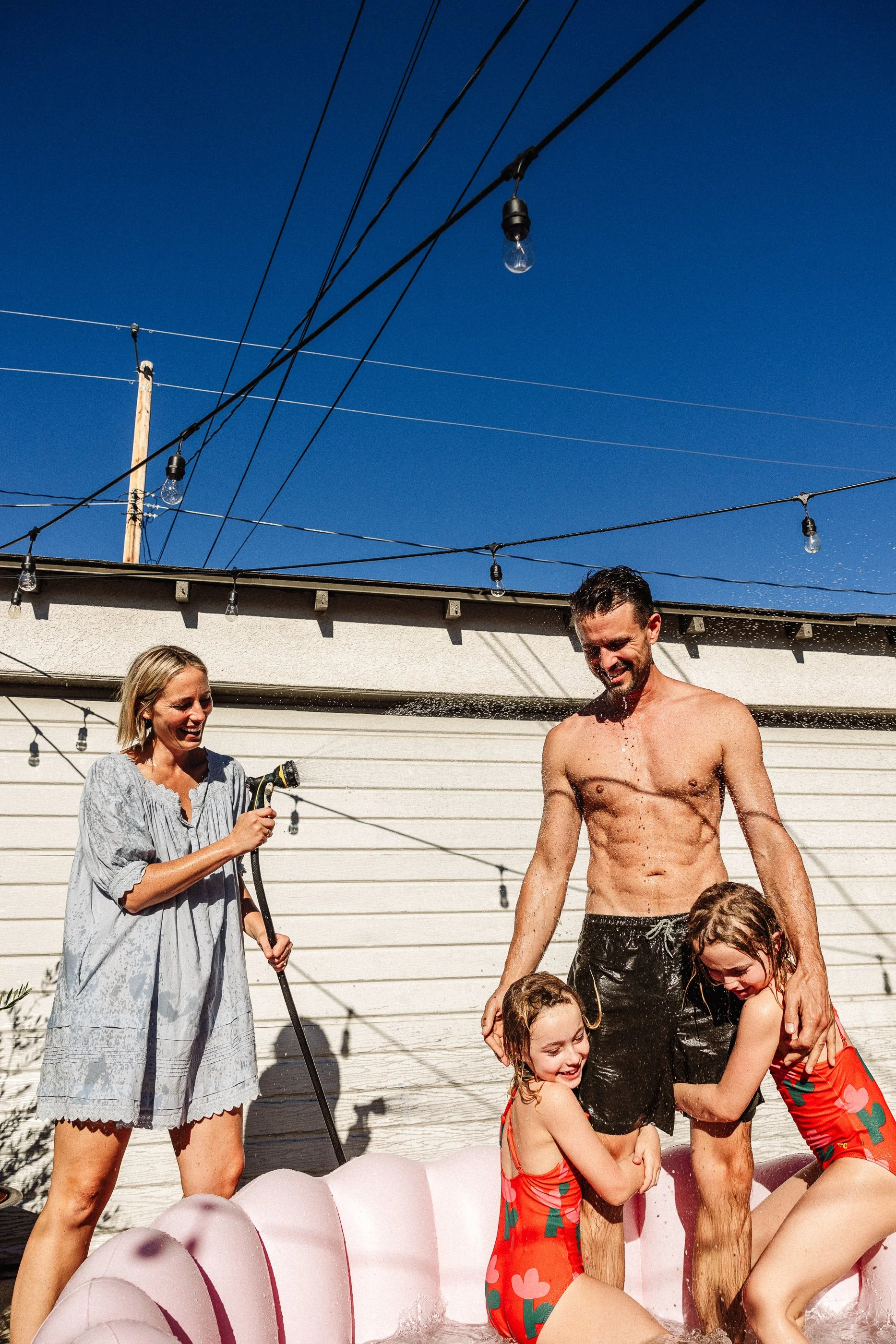 People enjoying a water activity in a backyard pool on a sunny day, with a woman holding a garden hose and children playing in the water. The background features a white garage door and string lights overhead.
