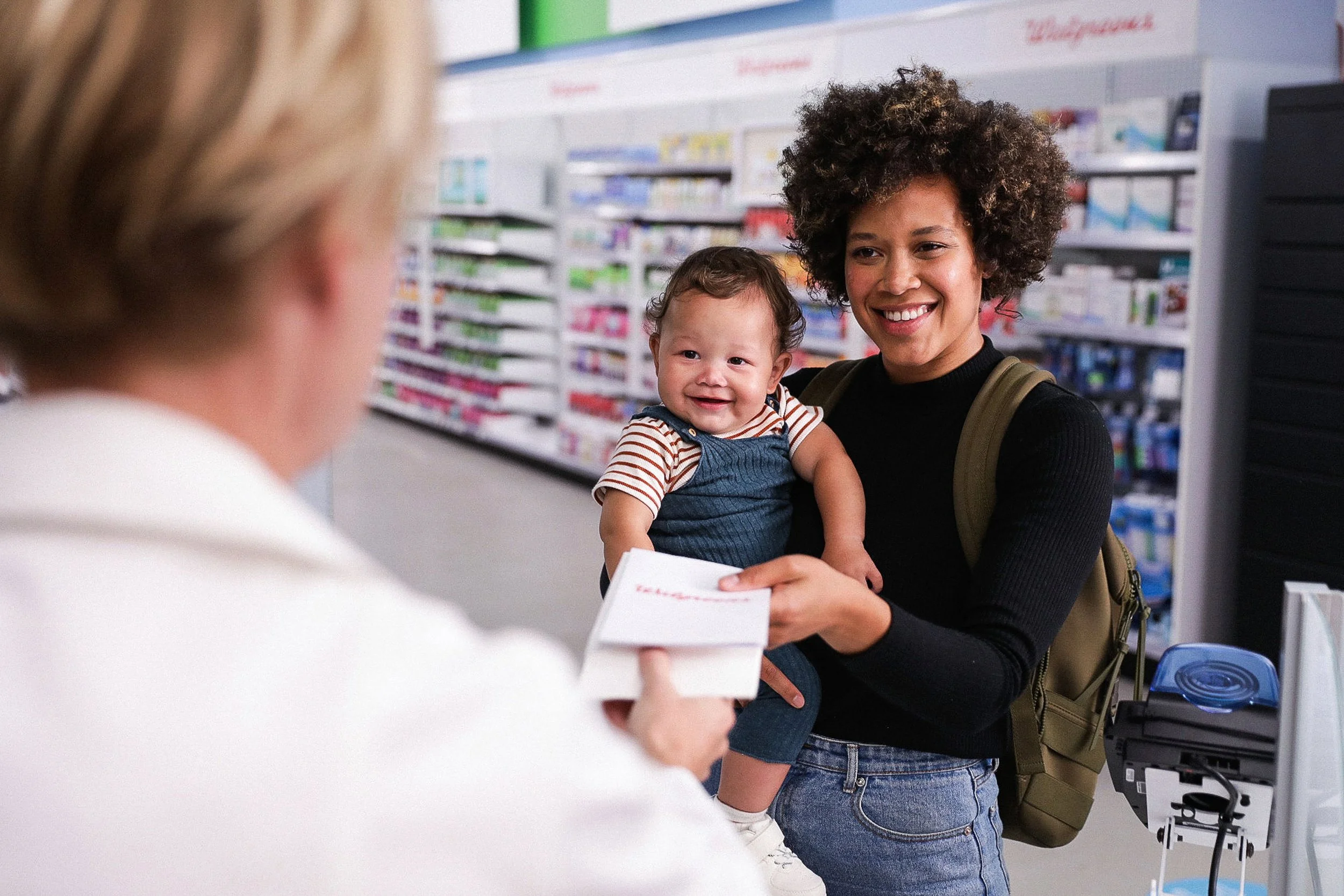 A woman with curly hair holding a baby and smiling at a person at a pharmacy counter. The person at the counter is holding a prescription.