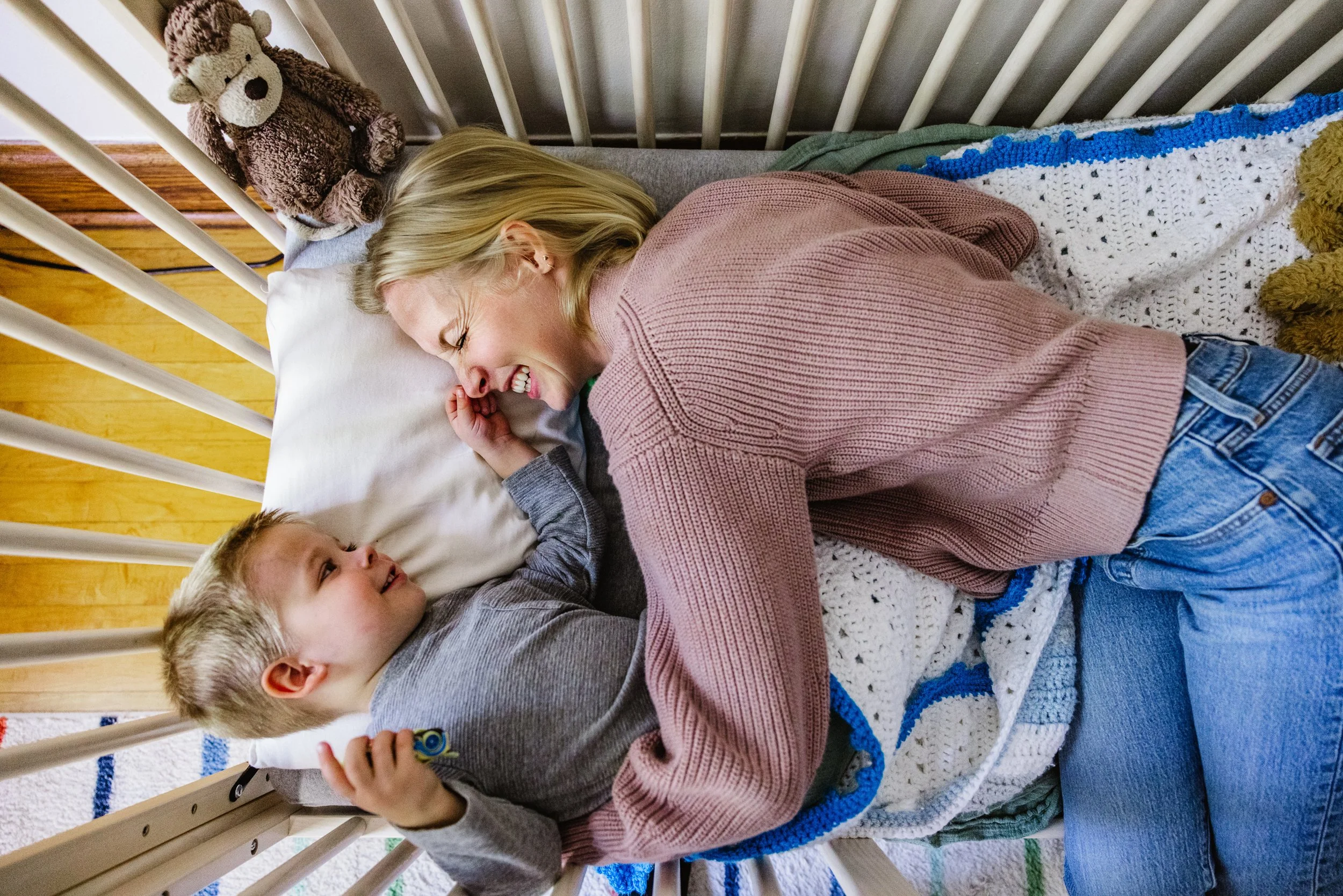 A woman and a young boy lying in bed, smiling and laughing together, with a teddy bear on the headboard and a crocheted blanket covering them.
