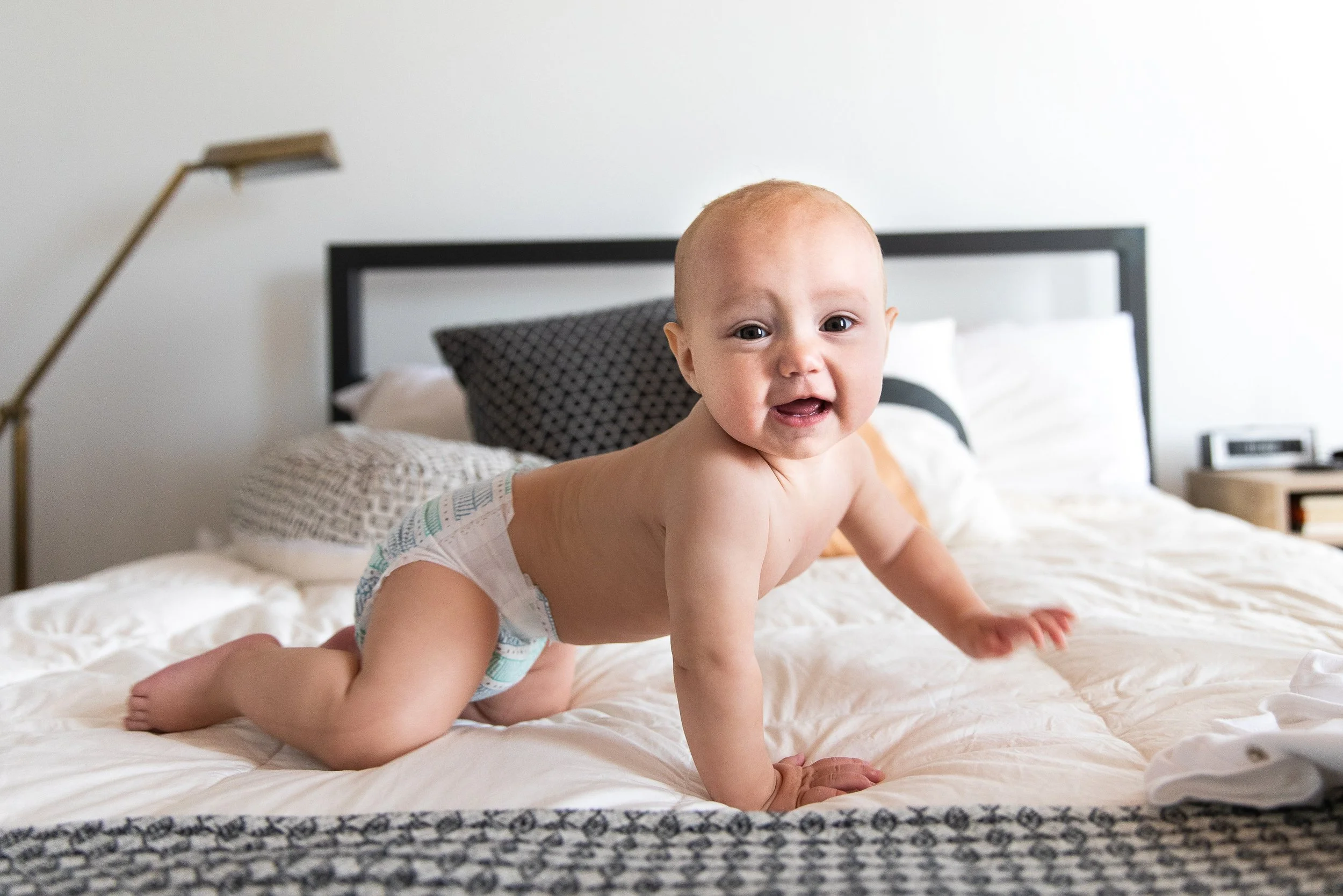 A smiling baby crawling on a bed with white sheets, a black headboard, and pillows, in a bright bedroom.