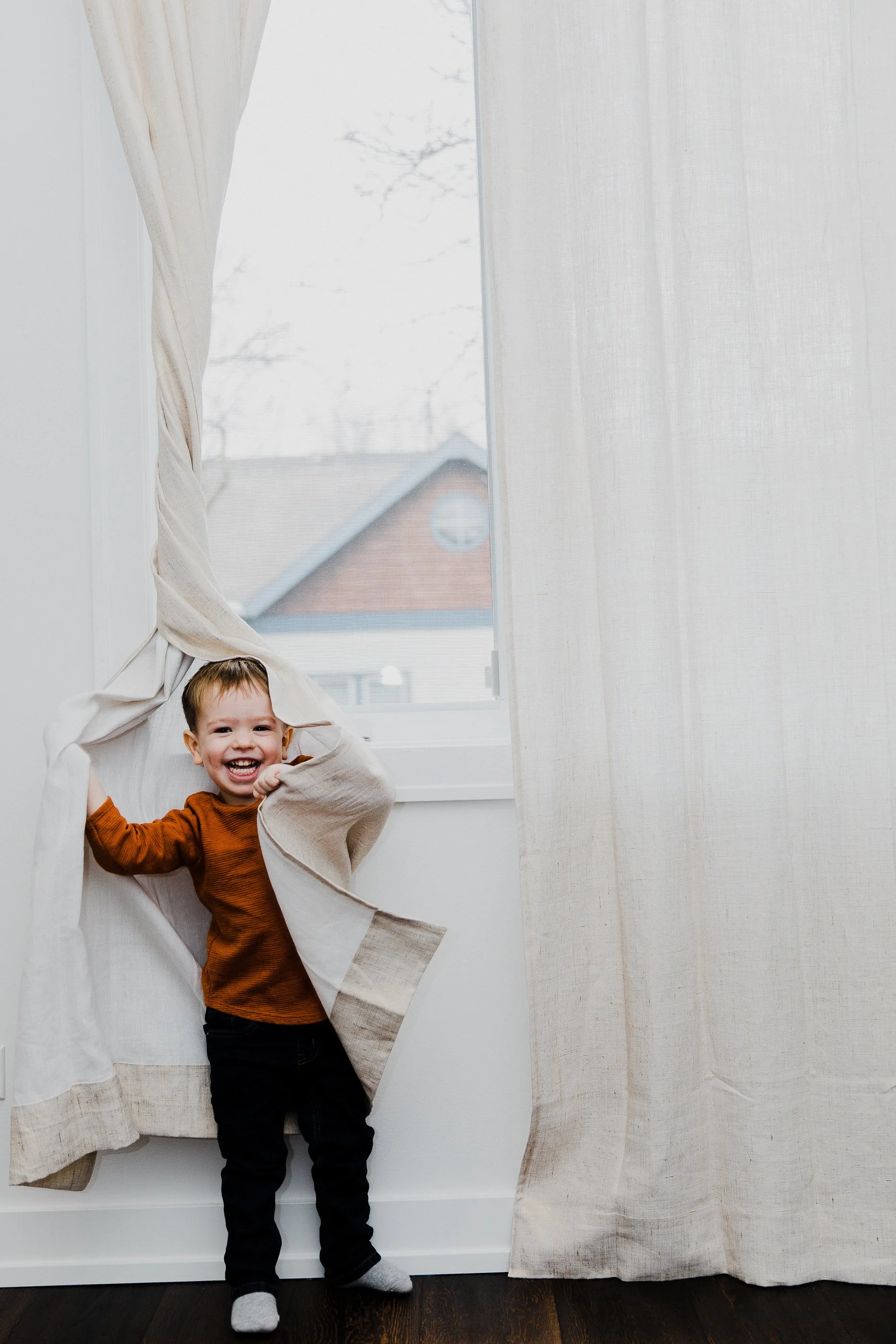 A young boy with brown hair, wearing a burnt orange shirt and black pants, is playing hide and seek behind a beige curtain, smiling and peeking out.