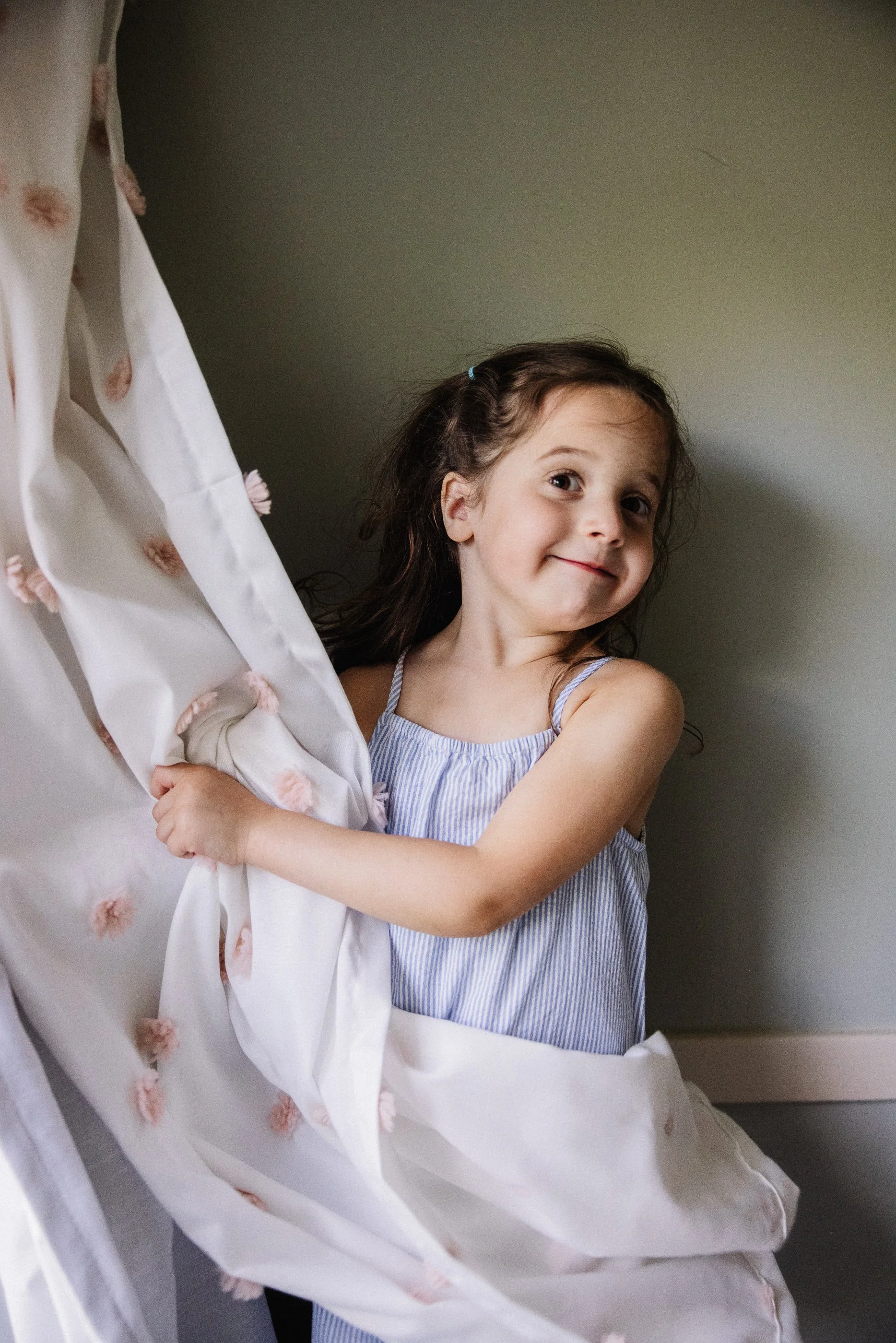 A young girl with brown hair and a blue hair tie, wearing a striped sleeveless dress, smiling and hugging a white blanket with pink floral embroidery.