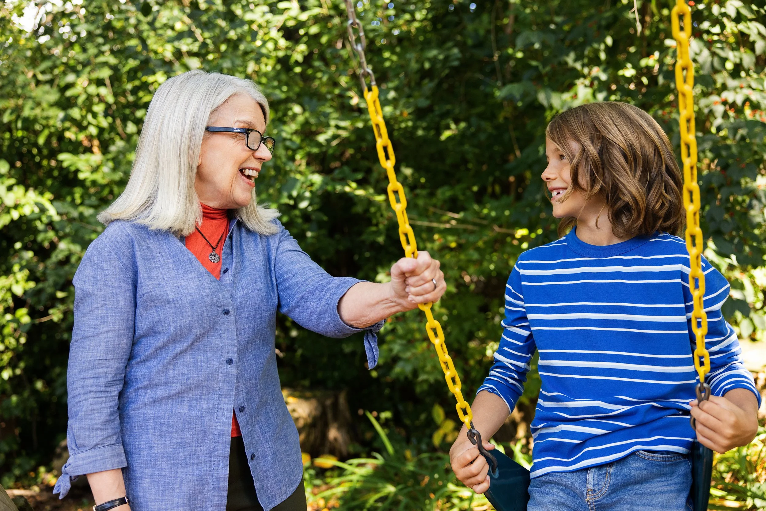 An elderly woman with glasses smiling and talking to a young boy with brown hair on a swing set in a green outdoor park.