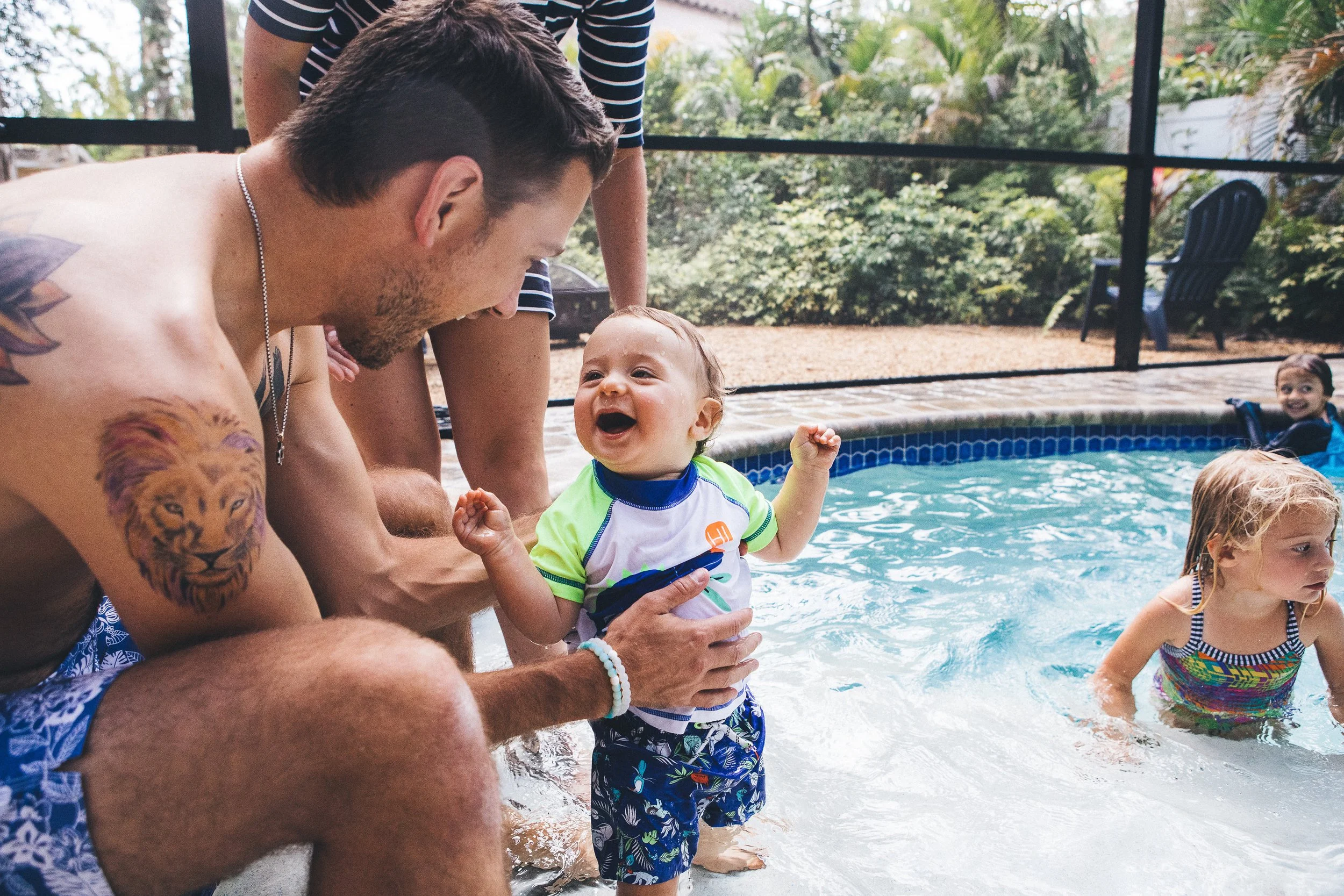 People, including children, enjoying time in a swimming pool outdoors surrounded by greenery and trees.