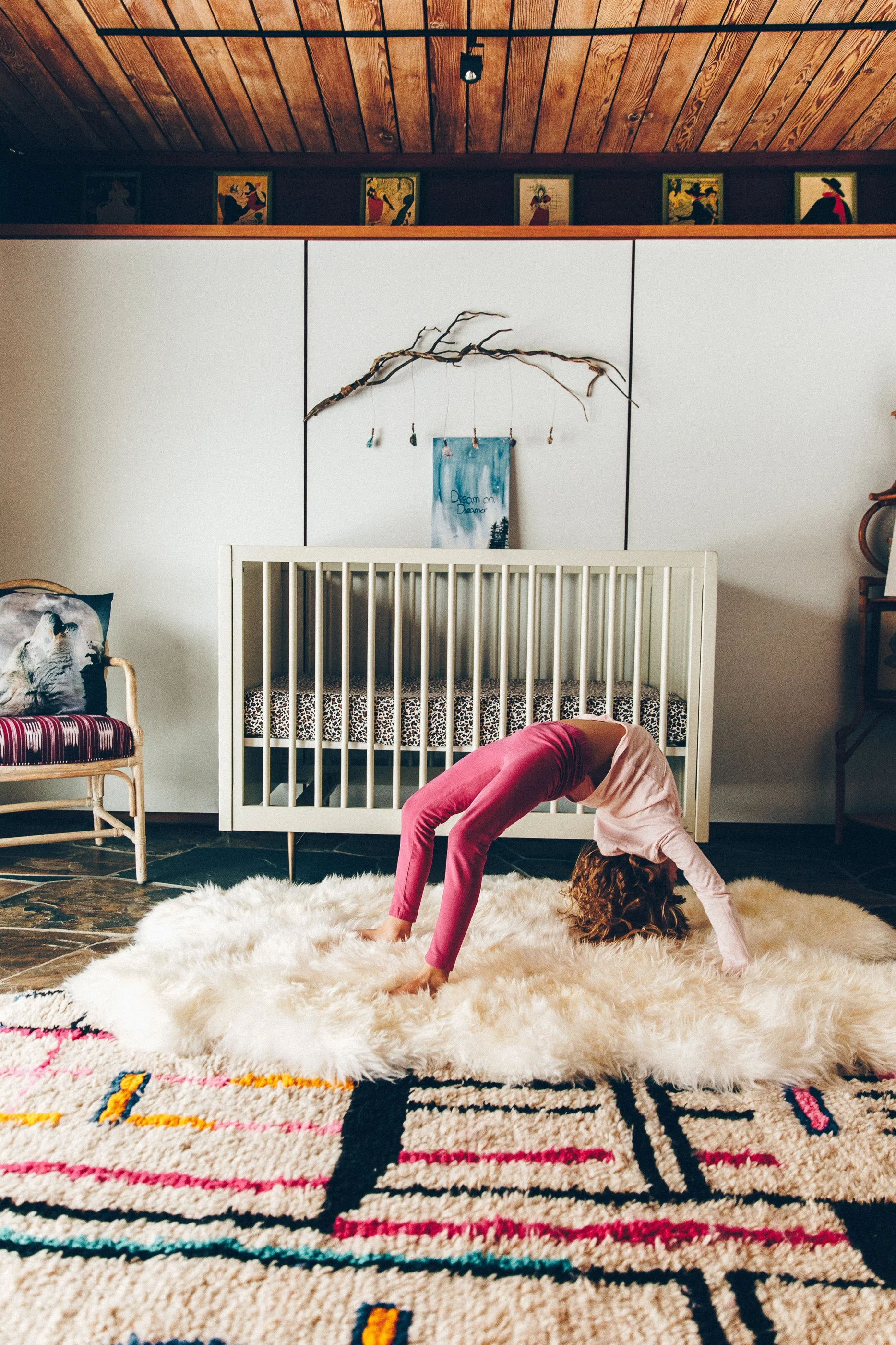 A girl with curly hair doing a downward dog yoga pose on a large cream-colored shaggy rug in a room with wooden ceiling and white walls. There is a patterned rug on the floor, a crib, a chair with a wolf print pillow, and framed artwork on the wall.