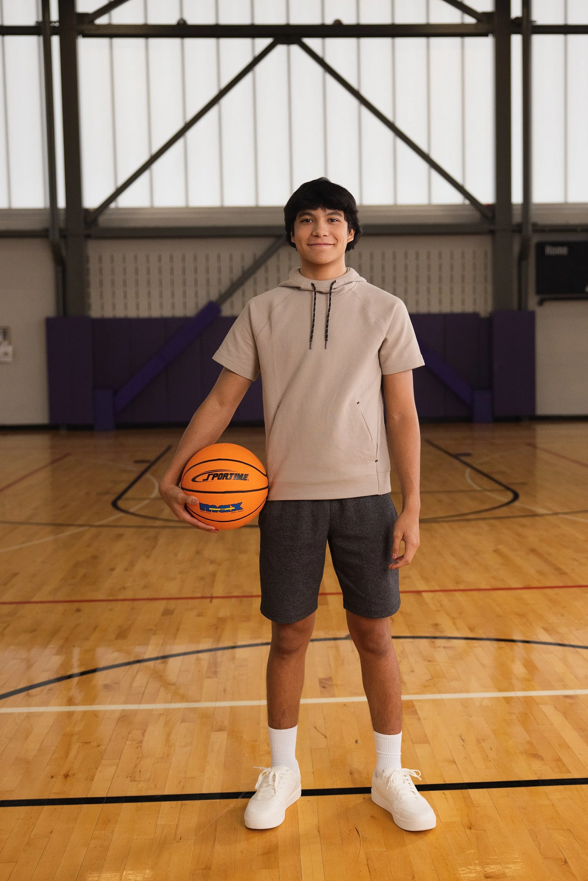 A young man stands in a basketball gymnasium holding a basketball with a smile. He wears a beige hoodie, dark shorts, white socks, and white sneakers. The gym has a wooden floor, and purple padded walls with a high ceiling featuring metal beams.