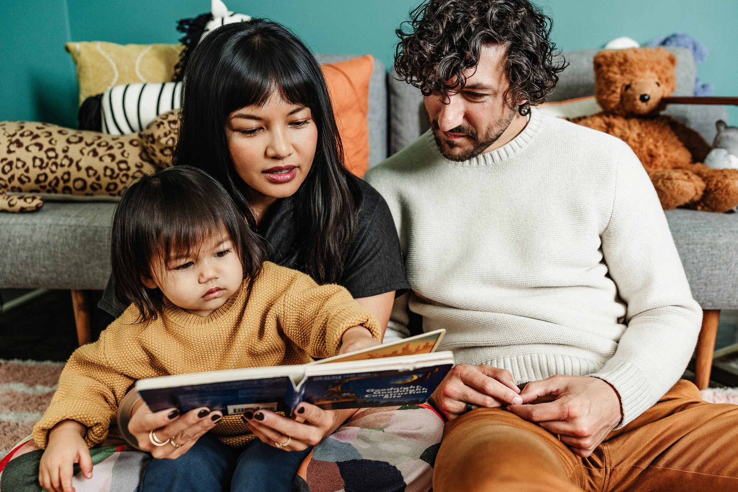 A family of three, a man with curly hair, a woman with straight black hair, and a young girl with straight hair, sitting on the floor together. They are looking at a book the woman is holding, in a cozy room with colorful pillows and teddy bears in t
