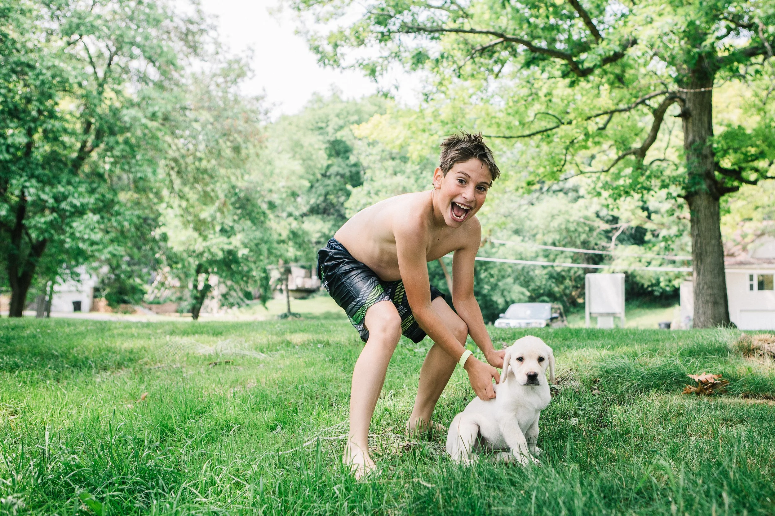 A young boy with short brown hair playing in a park with a light-colored puppy. The boy is smiling and wearing black swim shorts with green and yellow accents. The park has green grass and tall trees with lush foliage, and there are houses and cars v