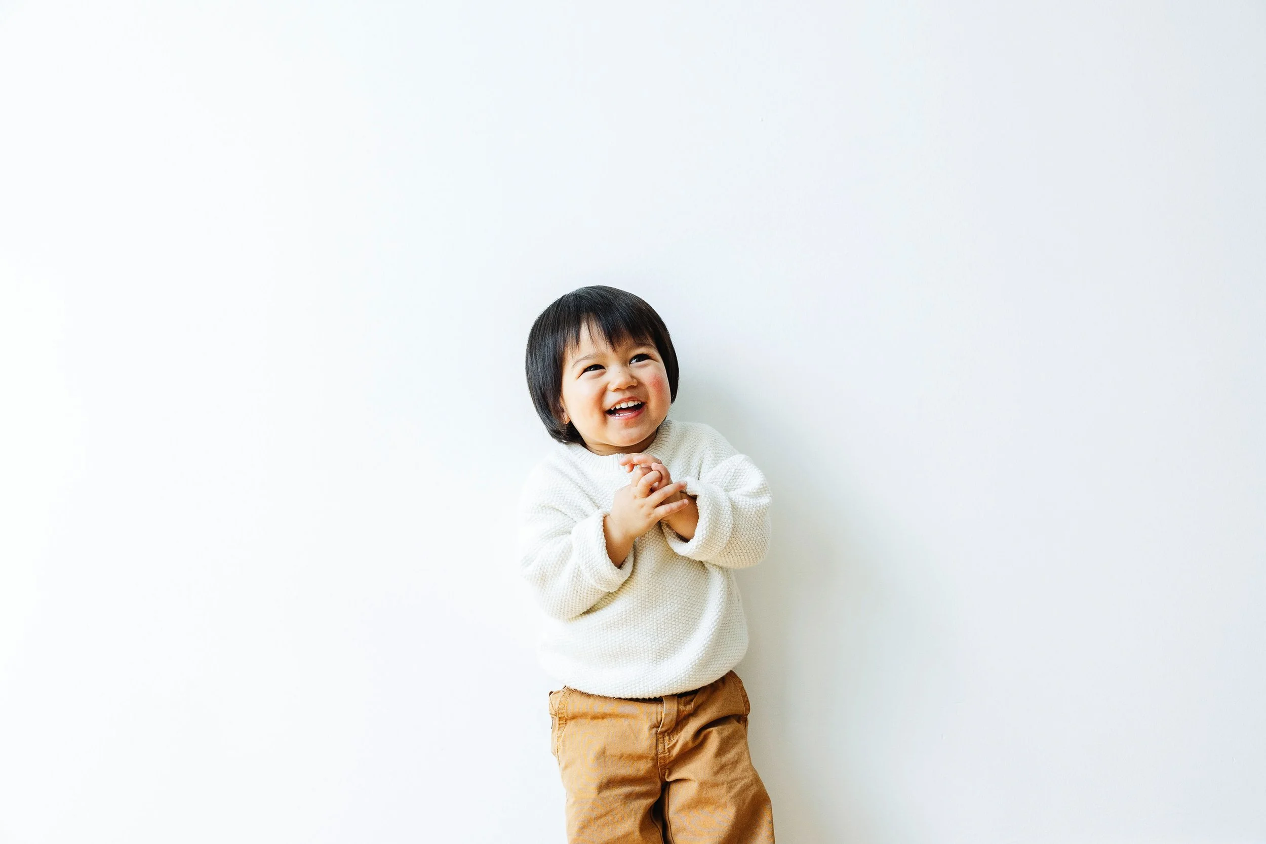 A young boy with black hair wearing a cream sweater and brown pants, smiling and clapping his hands while standing against a plain white wall.
