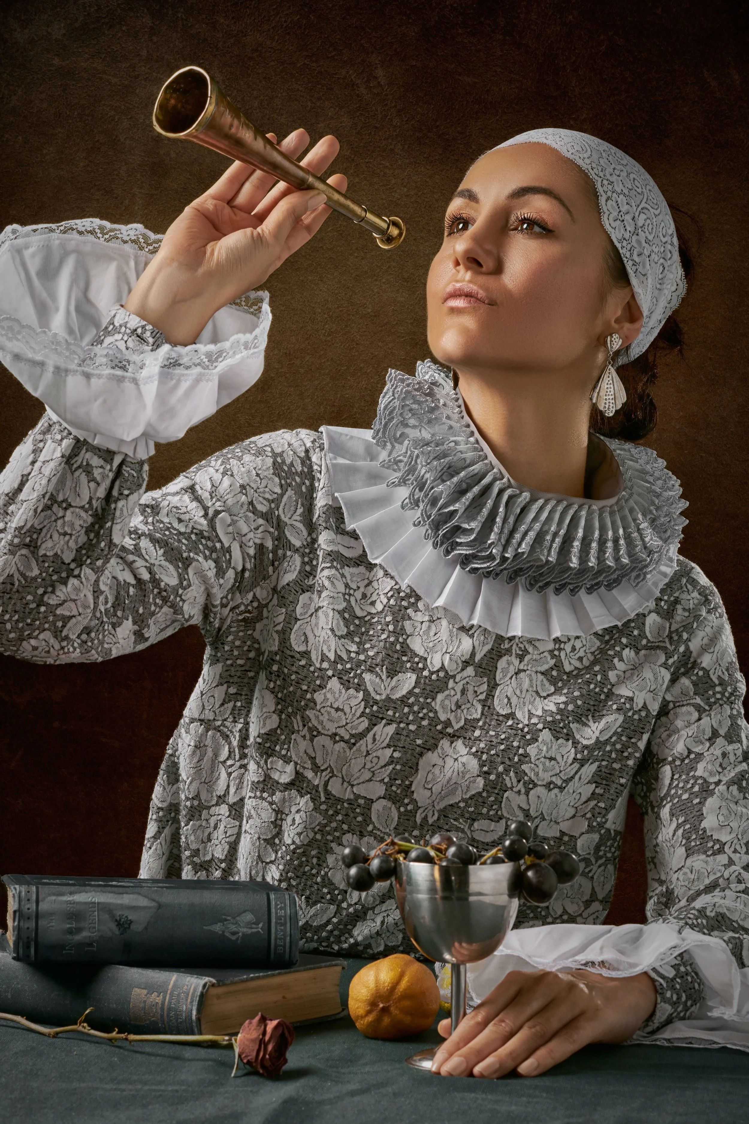 A woman dressed in an ornate, grey and white lace Renaissance-style gown and a lace head covering, holding an antique brass trumpet near her face, with a serious expression. She is seated at a table with open books, a silver goblet filled with grapes, a lemon, a withered rose, and a rolled-up document.